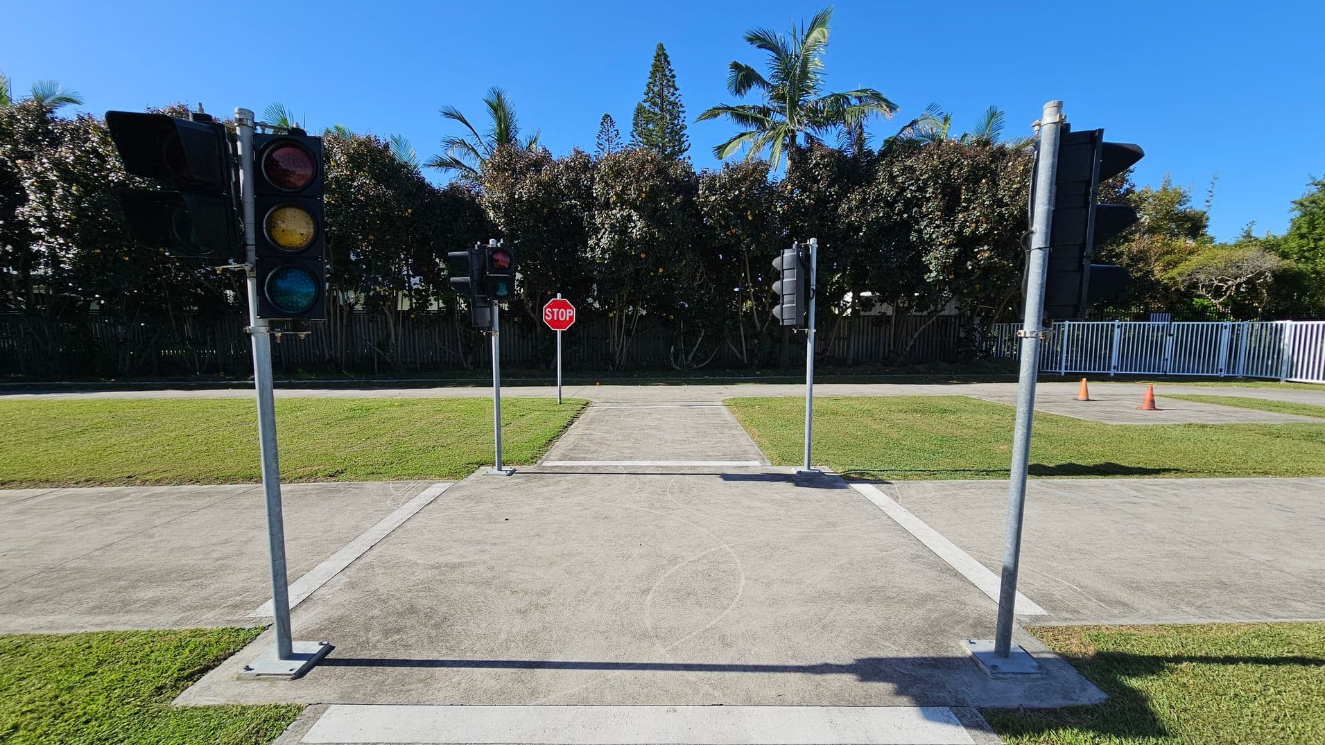 A small intersection with traffic lights on both sides and a stop sign in the centre. The ground is paved with concrete, surrounded by grass. In the background, there are tall trees and a white fence on the right. Two orange traffic cones are visible near the fence. The sky is clear and blue.