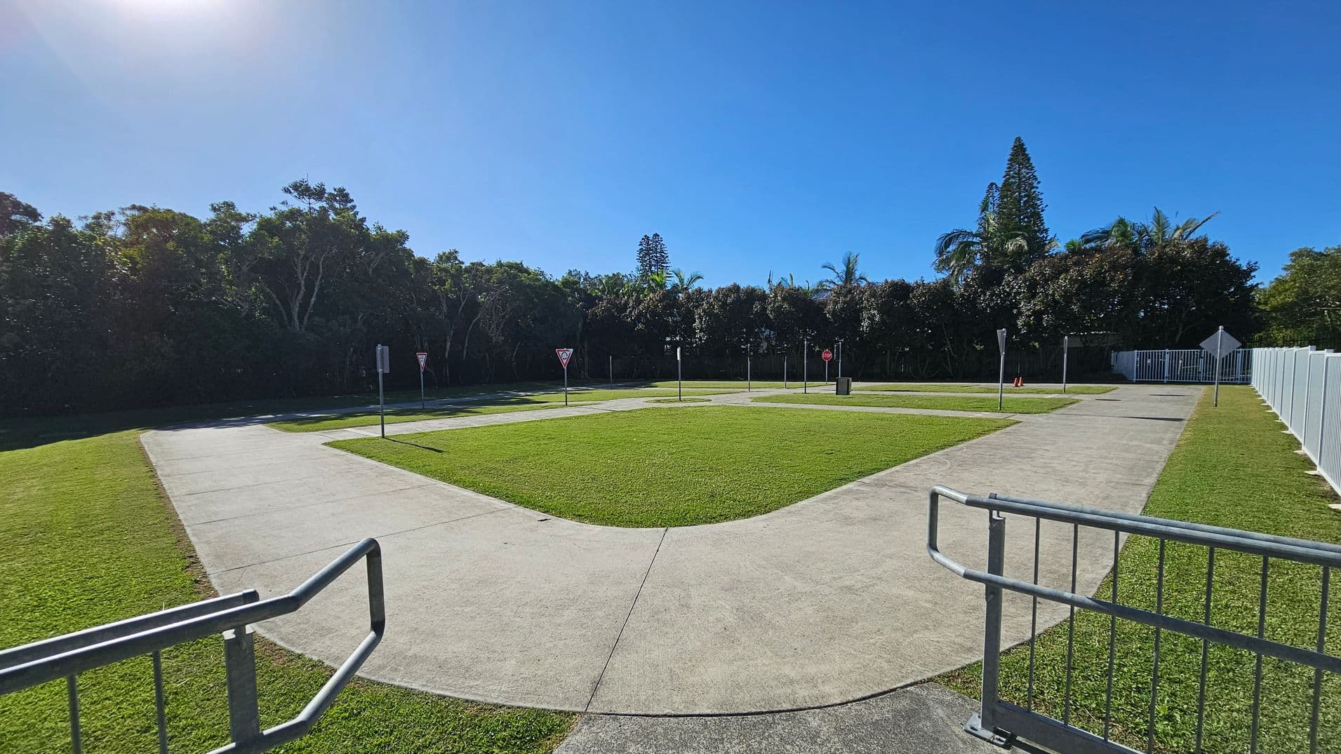 A small, fenced-in area with a paved path forming a loop around a grassy centre. Several traffic signs, including yield and stop signs, are placed along the path. The foreground features a metal railing. In the background, dense trees and tall coniferous trees are visible, with a clear blue sky above. The ground is covered with well-maintained grass.