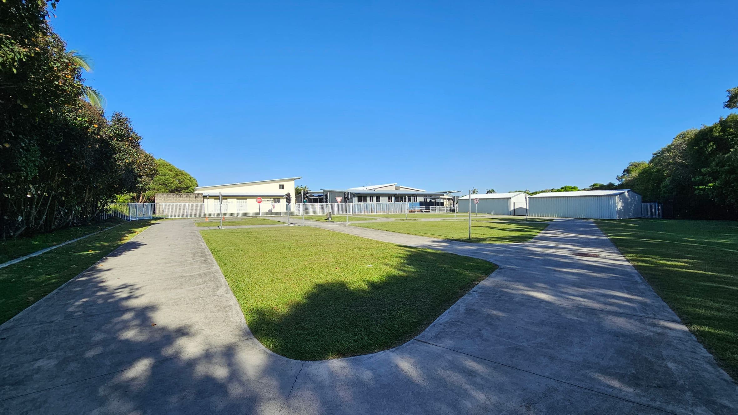 A paved path curves through a grassy area, leading towards a group of low, light-coloured buildings in the background. On the left, dense trees cast shadows on the path. On the right, more trees border the scene, with a white fence visible near the buildings. The sky is clear and blue.