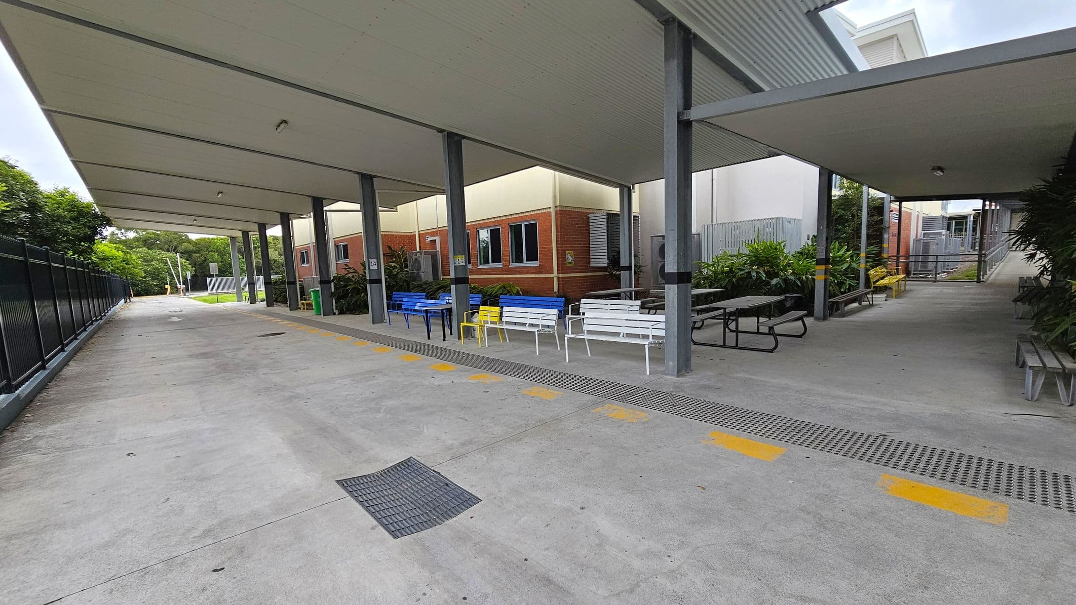 A covered walkway with a concrete floor and a metal ceiling supported by grey pillars. On the right, there are colourful benches in blue, yellow, and white, along with picnic tables. Behind the seating area, there are brick buildings with windows and some greenery. On the left, a black metal fence runs alongside the path, with trees visible in the background. Yellow markings are painted on the ground near the seating area.