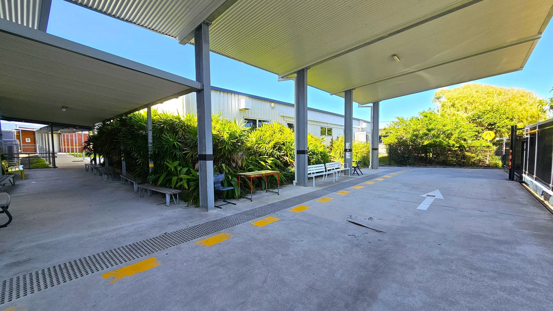 A covered outdoor walkway with a corrugated metal roof supported by metal beams. On the left, there are benches and a row of lush green plants. A white building with windows is visible behind the plants. On the right, a concrete path with a white arrow and yellow markings leads towards a fenced area with greenery. The ground is concrete, and the ceiling is metal.