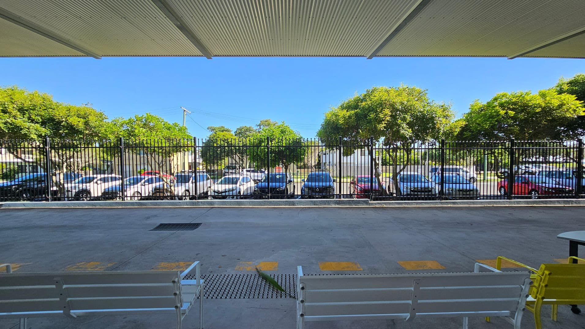 A covered area with a corrugated metal ceiling and a concrete floor. In the foreground, there are white benches and a yellow chair. Beyond a black metal fence, several parked cars are visible, including white, blue, and red vehicles. In the background, there are green trees and a clear blue sky.