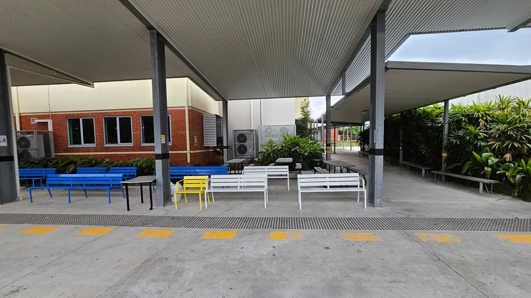 A covered outdoor area with a corrugated metal roof supported by metal beams. On the left, there are blue benches against a brick wall with windows and air conditioning units. In the centre, white benches and a yellow chair are arranged around tables. On the right, more benches are near lush green plants. The floor is concrete with yellow markings, and the ceiling is made of corrugated metal.