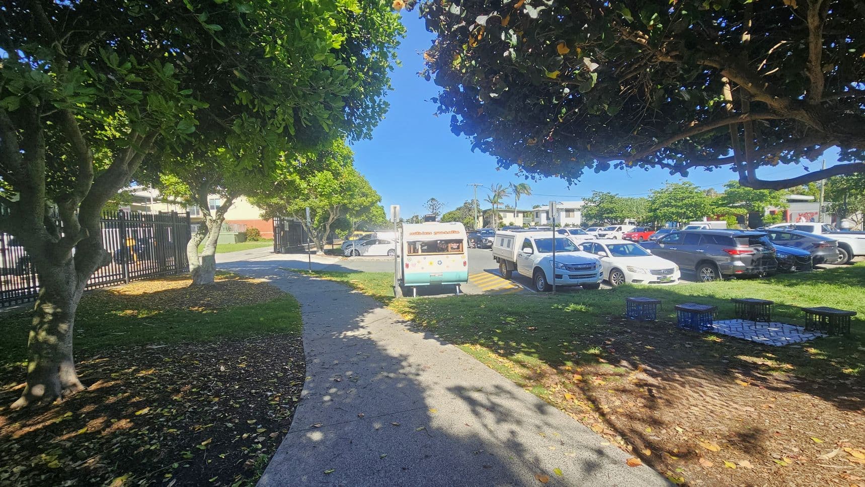 A pathway curves through a park area, bordered by trees with dense green foliage. On the left, a black metal fence runs alongside the path. In the centre, a small ice cream van is parked near the road. To the right, several parked cars are visible in a parking area. The ground is a mix of grass and mulch, with some fallen leaves. Milk crates are arranged on the grass, possibly as makeshift seating.