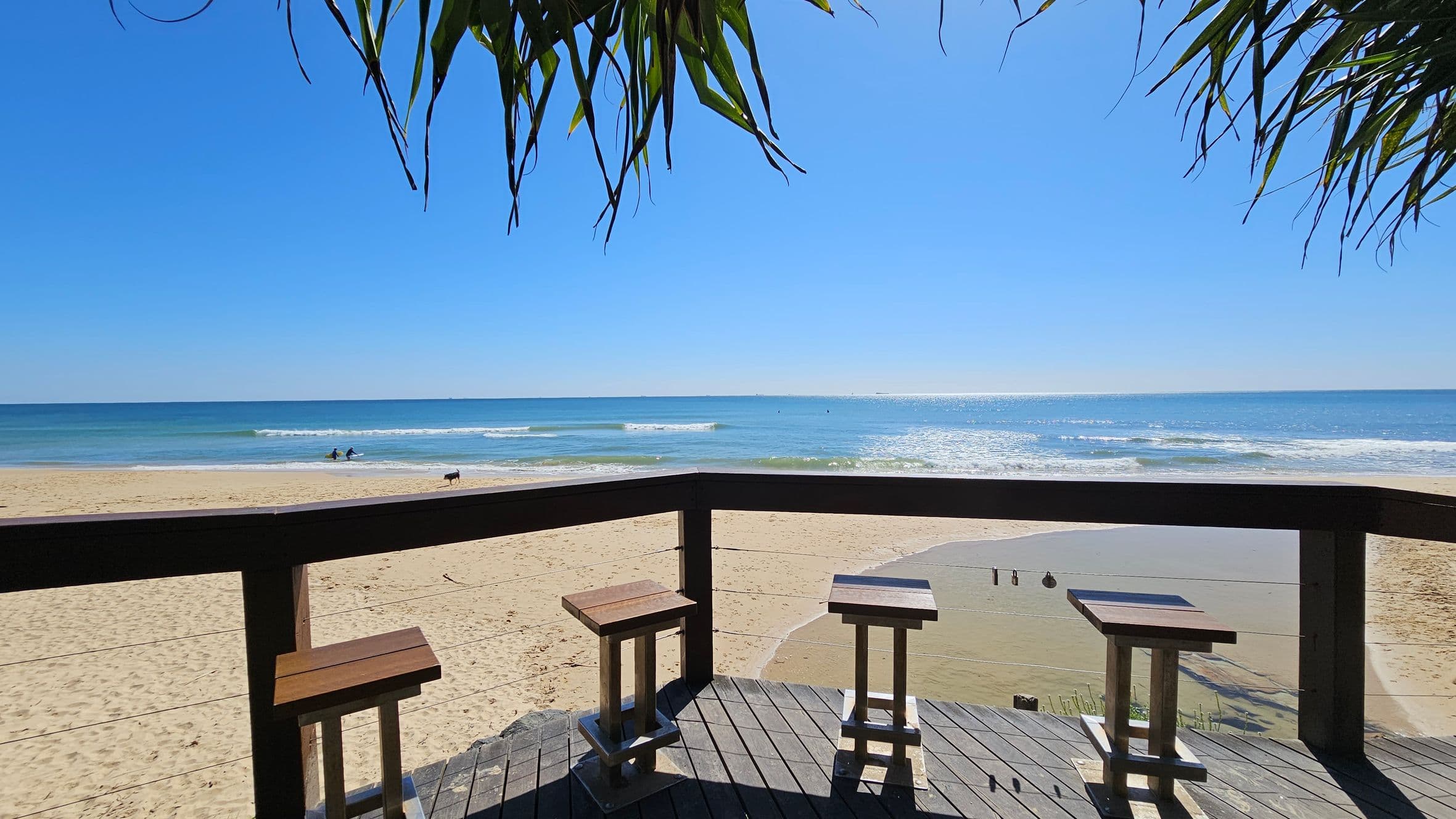 Wooden stools are arranged on a deck overlooking a sandy beach. The deck has a railing with a view of the ocean, where gentle waves meet the shore. In the background, two people are in the water, and a dog is walking along the sand. Overhead, leafy branches hang down, partially framing the scene. The sky is clear and blue.