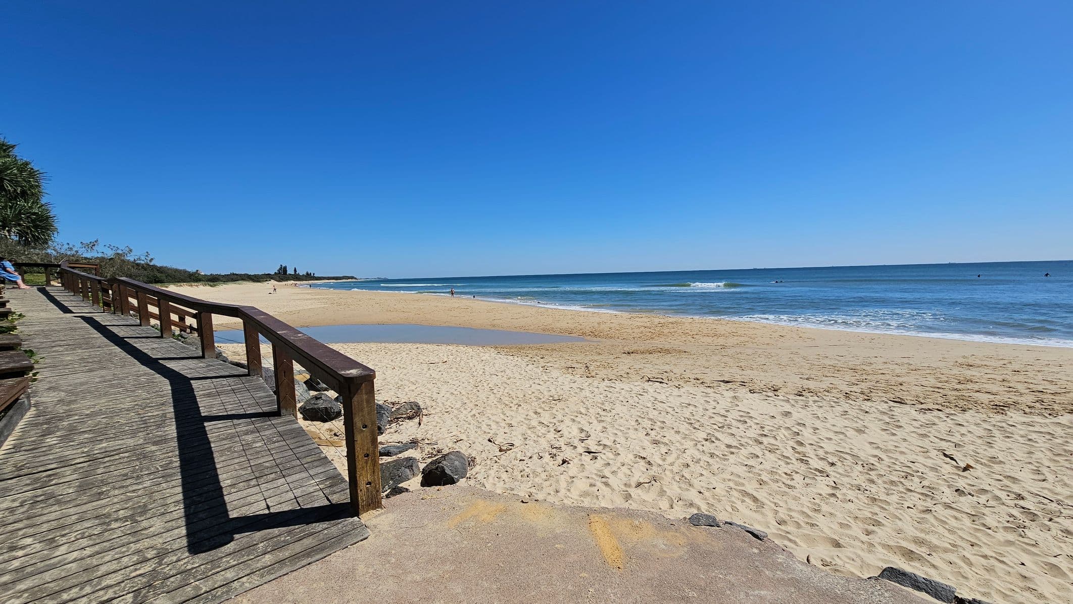 A wooden boardwalk with a railing runs along the left side, leading towards the background. On the right, a sandy beach stretches out to meet the ocean, with gentle waves lapping at the shore. In the distance, a few people are visible near the water. The foreground shows a sandy path with some rocks. Vegetation is visible on the far left, and the sky is clear and blue.