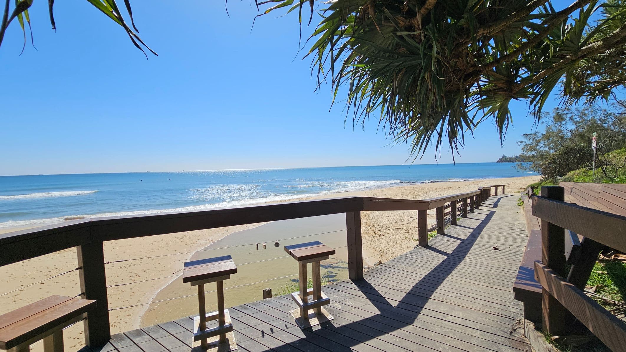 A wooden boardwalk runs along a sandy beach, with two stools positioned at a railing overlooking the ocean. The sea is calm with gentle waves. On the right, the boardwalk extends into the distance, bordered by greenery. Overhead, leafy branches hang down, partially shading the area. The beach stretches out to the left, meeting the blue water under a clear sky.