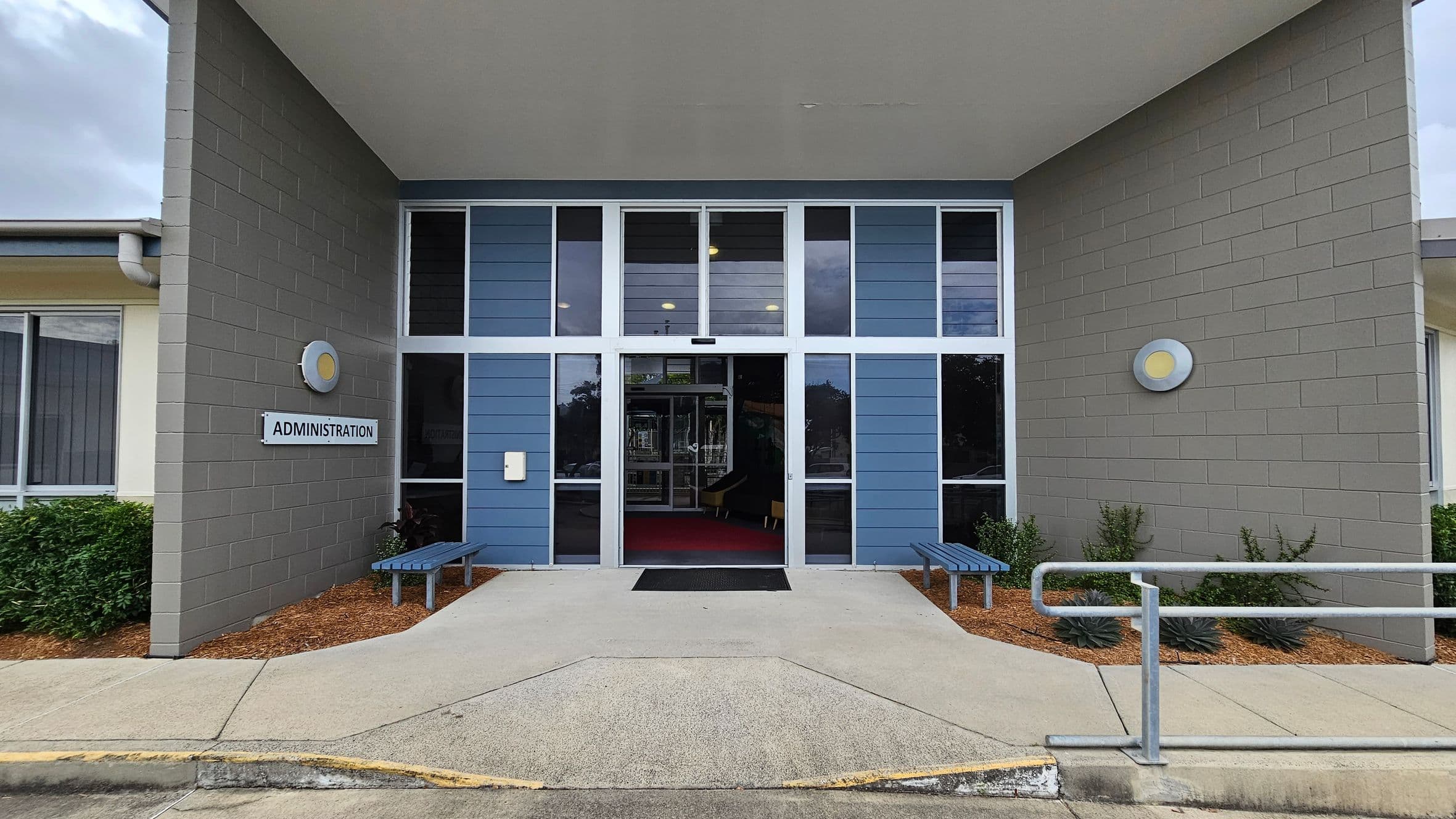 Entrance to a building with a sign reading "Administration" on the left wall. The facade features grey brick walls and blue panelling with large glass windows. Two circular lights are mounted on each side. A pathway leads to the entrance, flanked by two blue benches. A metal railing is on the right, and there are small shrubs planted along the walls. The ceiling is flat and extends over the entrance area.