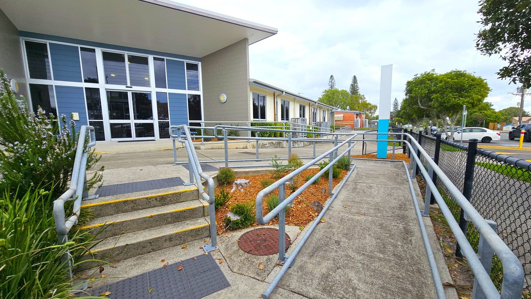 A building with large blue and grey windows on the left, featuring a ramp and steps with metal railings leading up to the entrance. The foreground has a landscaped area with small plants and mulch. On the right, a black chain-link fence runs alongside a pavement, with trees and parked cars visible in the background. The ground is concrete, and the ceiling of the entrance area is flat and white.