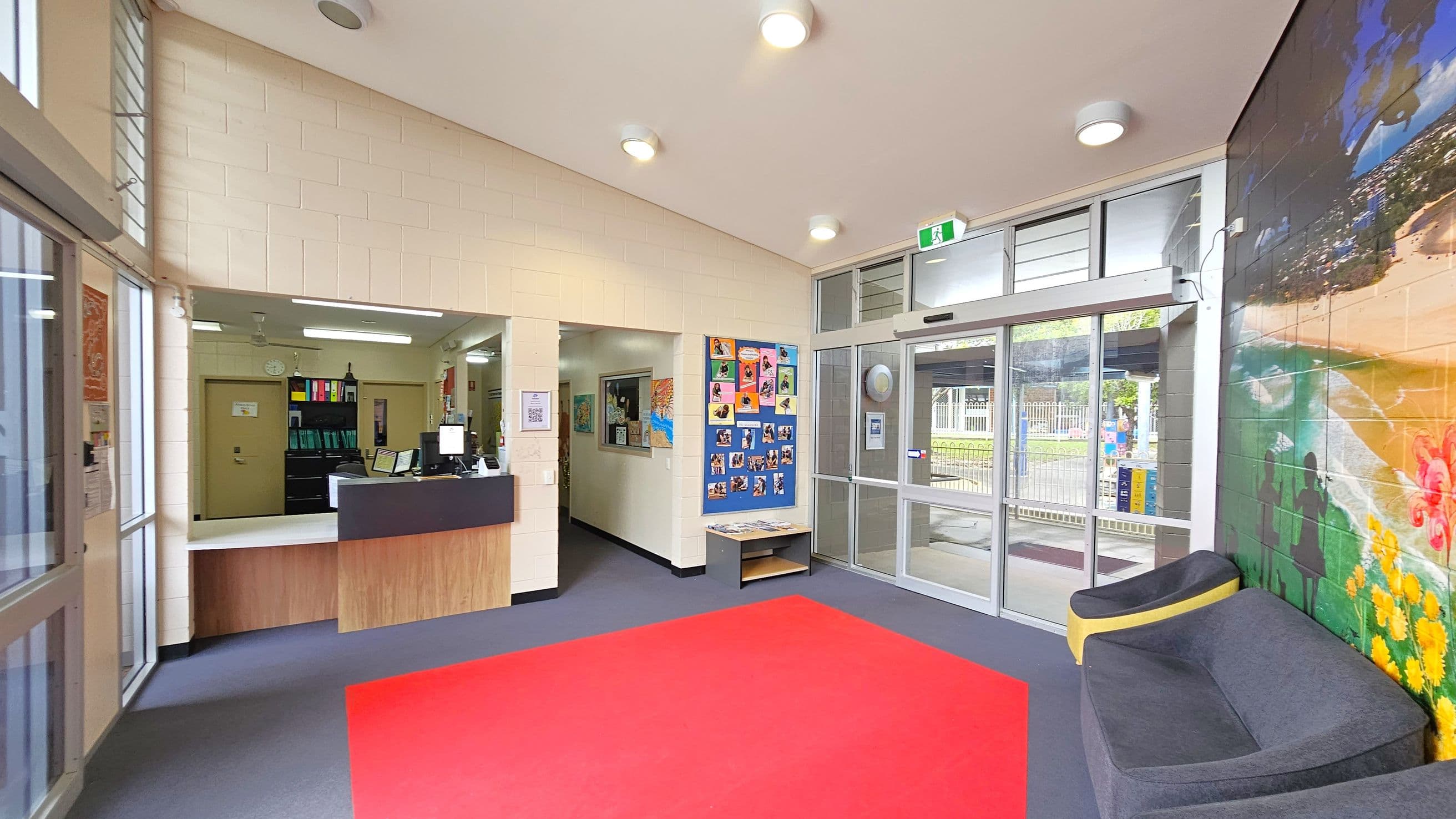A reception area with a counter on the left, featuring a computer and office supplies. Behind the counter, there are shelves with colourful binders. In the centre, a wall displays a bulletin board with photos and artwork. To the right, a glass door leads outside, with a mural of a beach scene on the adjacent wall. A grey and yellow seating area is positioned near the mural. The floor is covered with a red carpet, and the ceiling has several round lights.