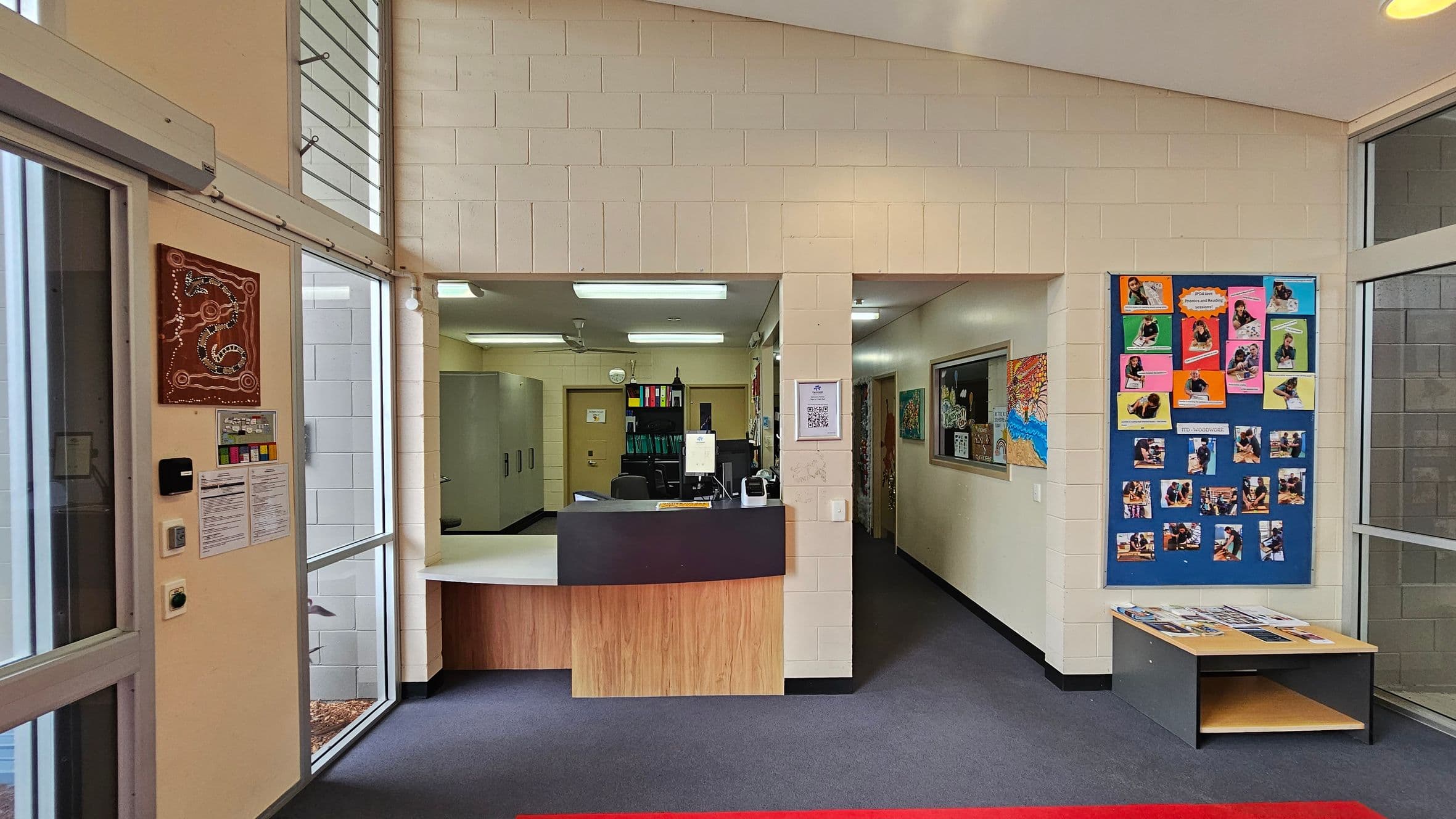 A reception area with a counter in the centre, featuring a computer and office supplies. To the left, there are large windows with a wall-mounted artwork and notices. On the right, a colourful bulletin board displays various photos and papers. Below the board is a small table with brochures. The floor is carpeted in dark grey, and the ceiling has fluorescent lights.