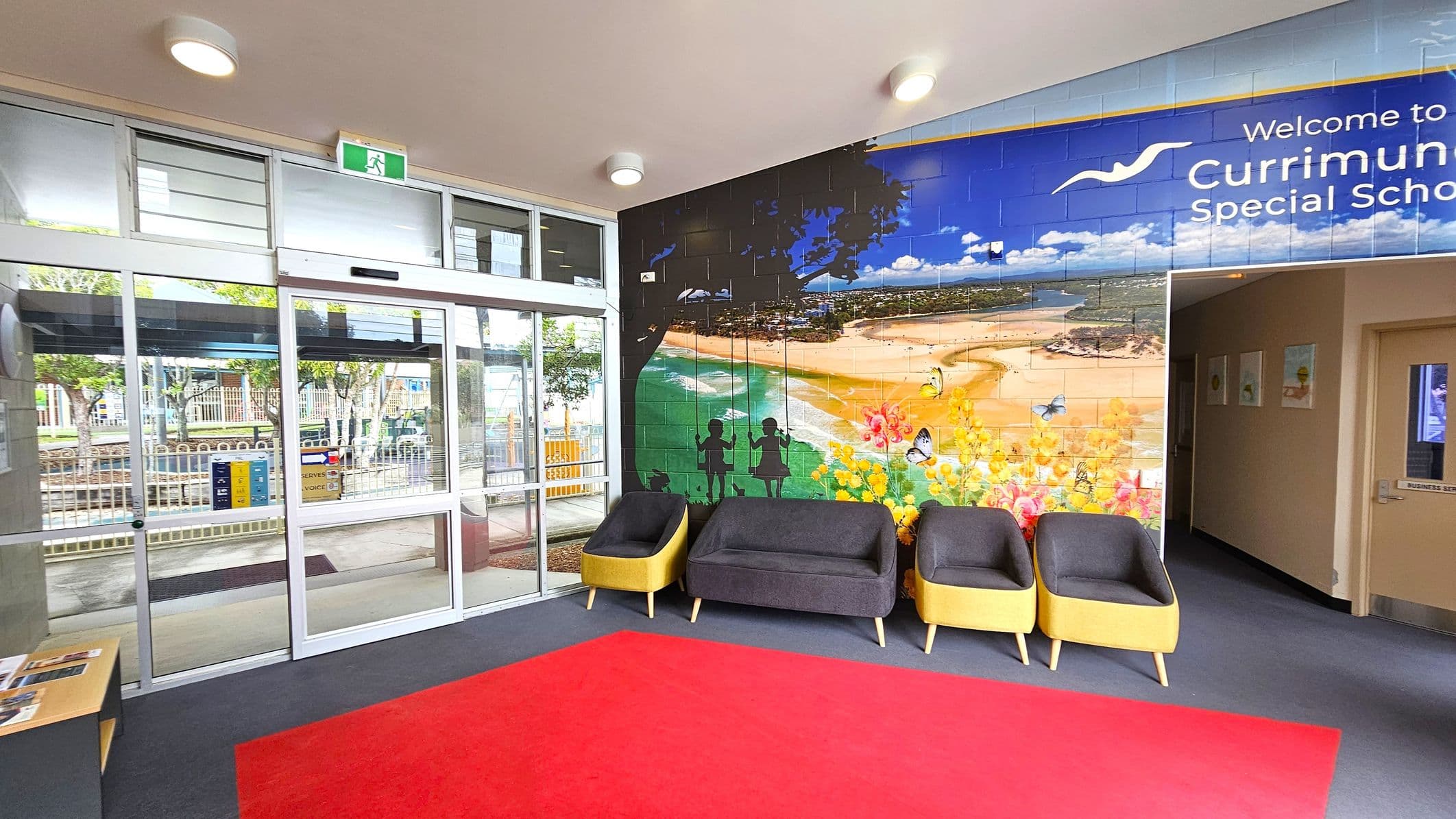 Entrance area with a glass sliding door on the left, leading outside. Above the door is an emergency exit sign. The right wall features a large mural depicting a beach scene with silhouettes of children on swings and colourful flowers. Below the mural are three chairs with grey cushions and yellow sides. The floor is covered with a red carpet, and the ceiling has round lights. A small table with brochures is near the door.