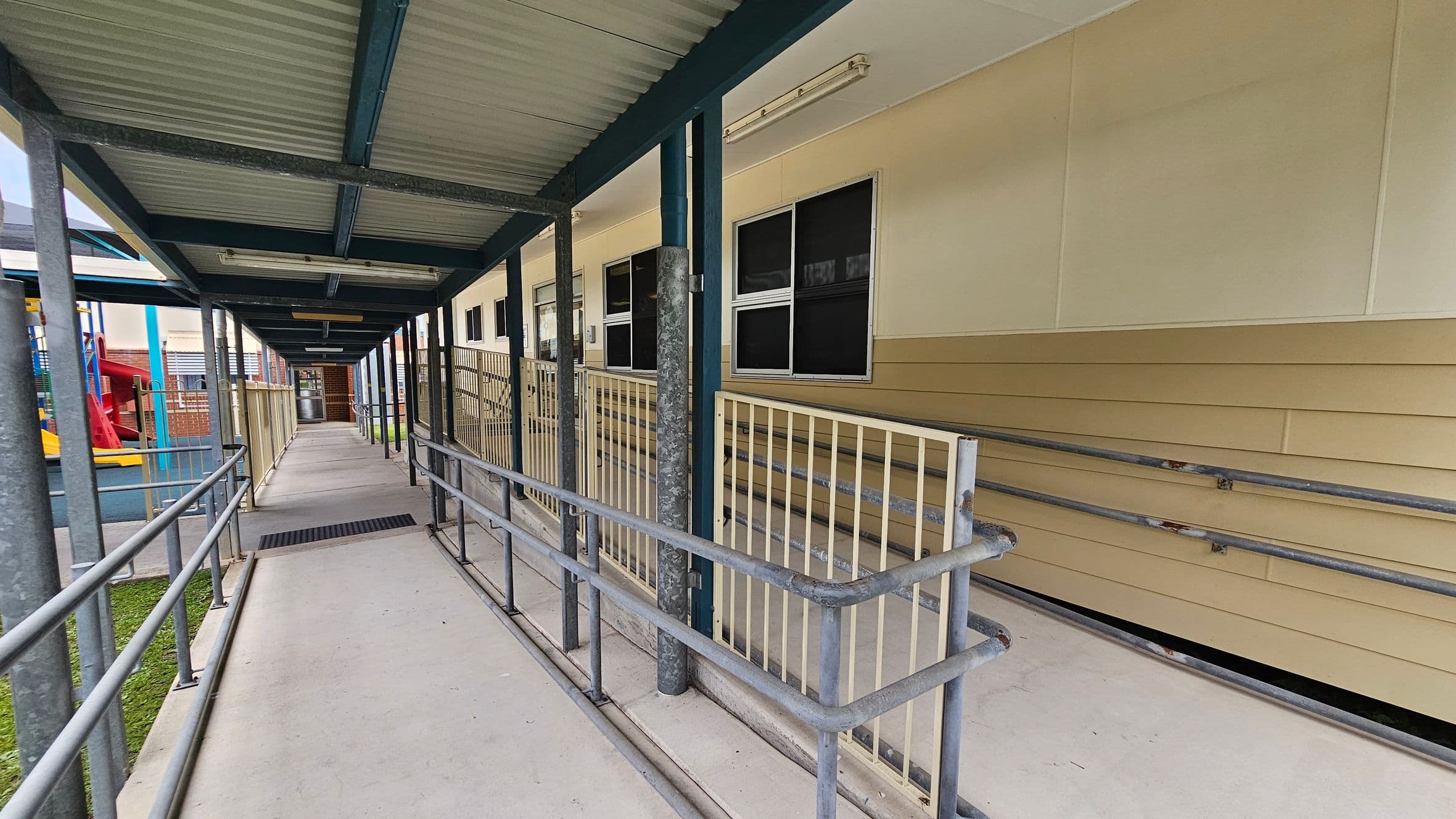 A covered walkway with a corrugated metal roof and metal railings on both sides. On the right, there is a beige building with several windows and a ramp leading up to it. On the left, a playground area is visible with a red slide and blue play structures. The floor is concrete, and the ceiling is supported by metal beams. The walkway extends into the distance, leading to a door at the far end.