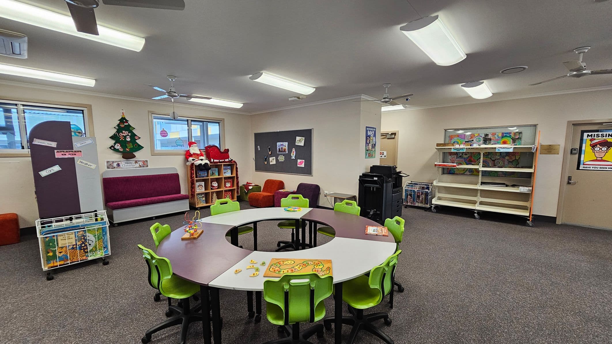 A classroom with a circular table in the centre surrounded by green chairs. On the table are educational toys and games. To the left, a purple bench sits beneath a window, with a Christmas tree decoration and a Santa figure on a shelf. The right side has a large bulletin board with artwork and a printer nearby. Shelves with books and a "Missing" poster are on the far right wall. The ceiling has fluorescent lights and fans, and the floor is carpeted.