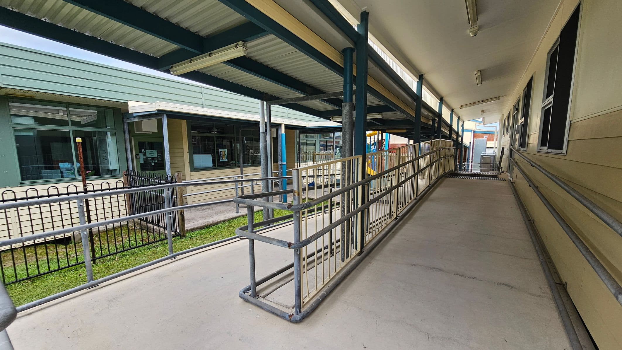 A covered walkway with a ramp and metal railings on both sides. On the right, there is a beige building with several windows. On the left, a green building with large windows and a black metal fence. The floor is concrete, and the ceiling is made of corrugated metal with fluorescent lights. In the background, there are more structures and a glimpse of a playground with colourful equipment.