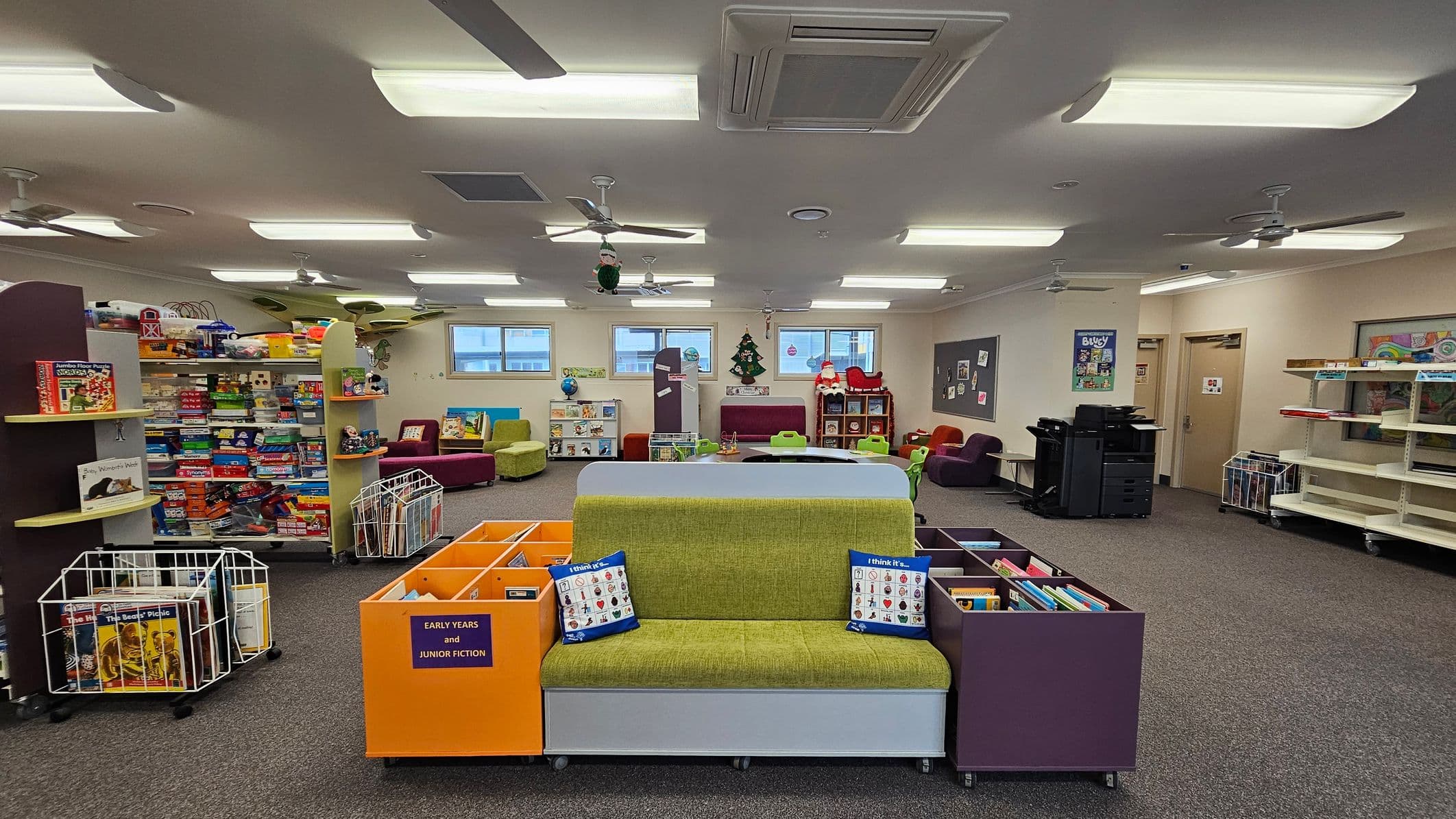 A spacious room with a carpeted floor and a ceiling fitted with fluorescent lights and ceiling fans. In the foreground, a green cushioned bench is flanked by colourful book storage units labelled "Early Years and Junior Fiction." To the left, shelves are filled with board games and educational toys. The background features windows, a Christmas tree decoration, and a Santa figure. On the right, there are printers and empty shelves. The room has a bright and organised appearance.