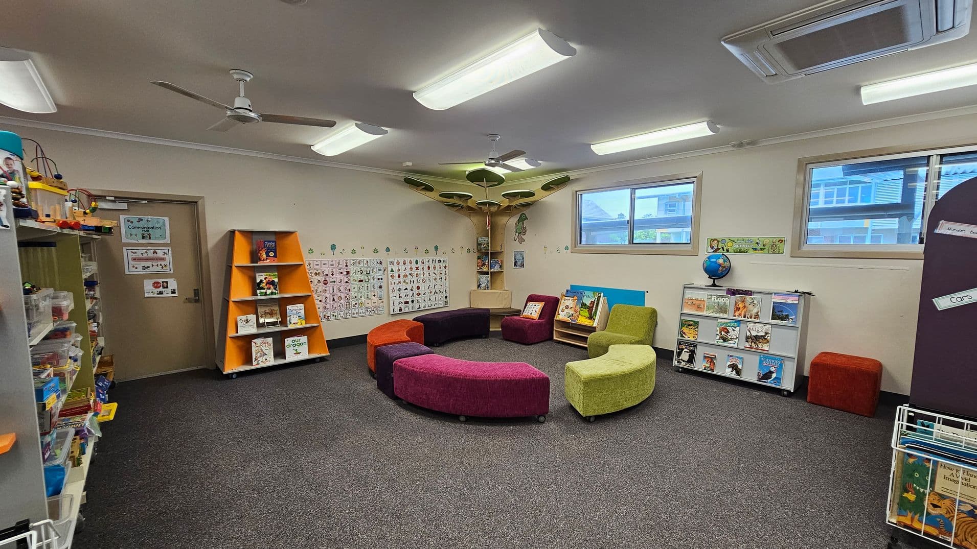 A classroom with a grey carpeted floor and a ceiling with fluorescent lights and ceiling fans. In the centre, there are colourful, curved seating pieces arranged in a circle. On the left, shelves hold various educational materials and toys. The back wall features a door and an orange triangular bookshelf with books. The right side has windows, a globe on a low shelf with books, and a small book display. The walls are decorated with educational posters and artwork.