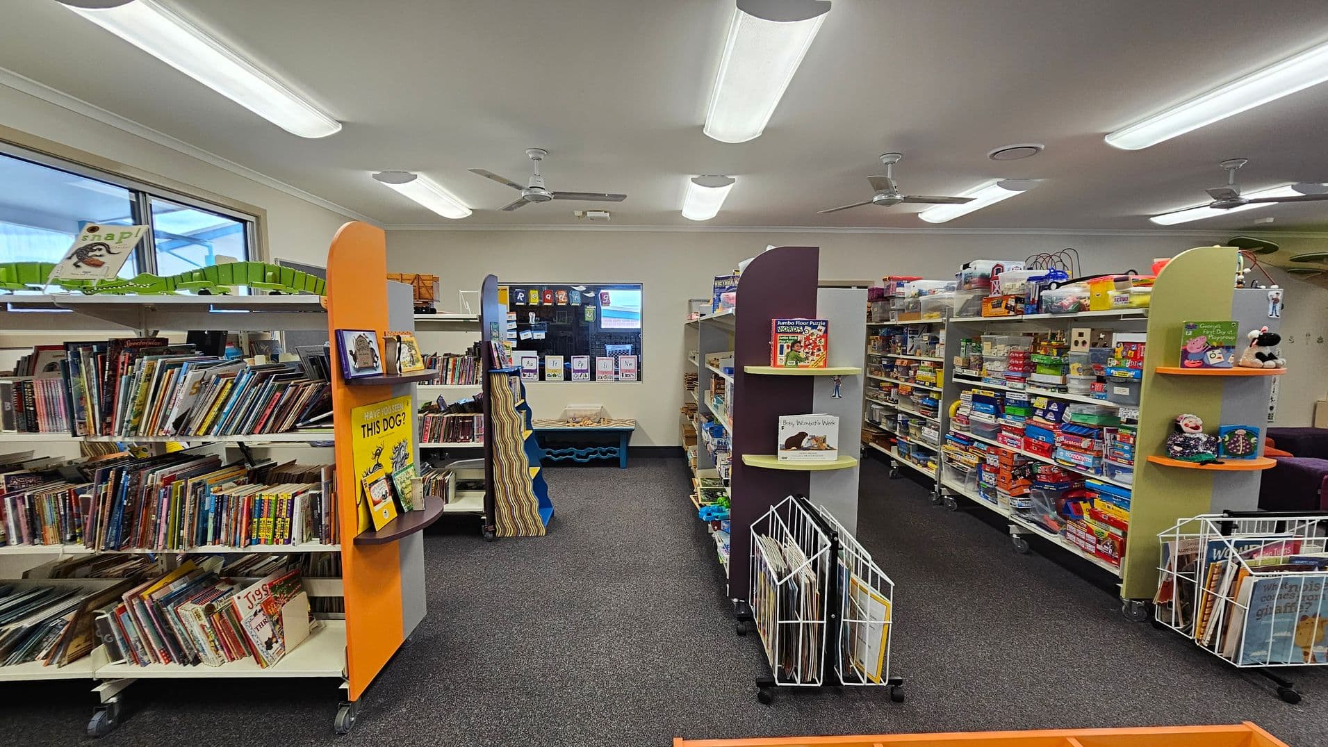 Bookshelves filled with children's books and games line both sides of a library room. On the left, shelves are packed with colourful books, while the right side features games and educational toys. In the centre, a small display shelf holds a few featured books. The floor is carpeted in grey, and the ceiling has fluorescent lights and ceiling fans. A window at the back shows a glimpse of the outside, and there are educational posters on the walls.