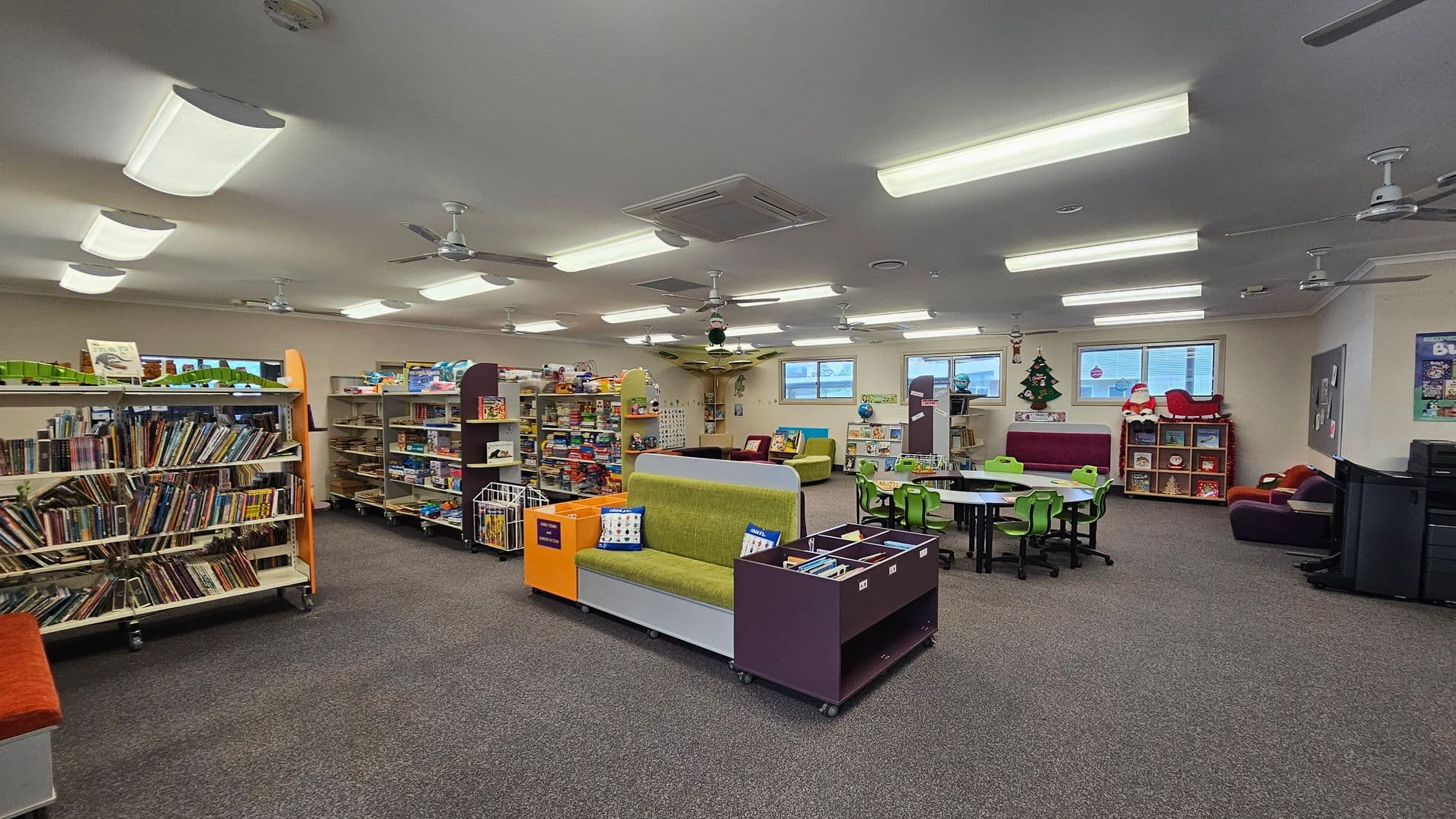 A spacious library room with grey carpeted flooring and a ceiling fitted with fluorescent lights and ceiling fans. On the left, there are bookshelves filled with books. In the centre, a green sofa with colourful cushions is flanked by storage units. To the right, there are tables with green chairs and a small seating area with plush toys. The background features windows and festive decorations, including a Christmas tree and Santa figure.