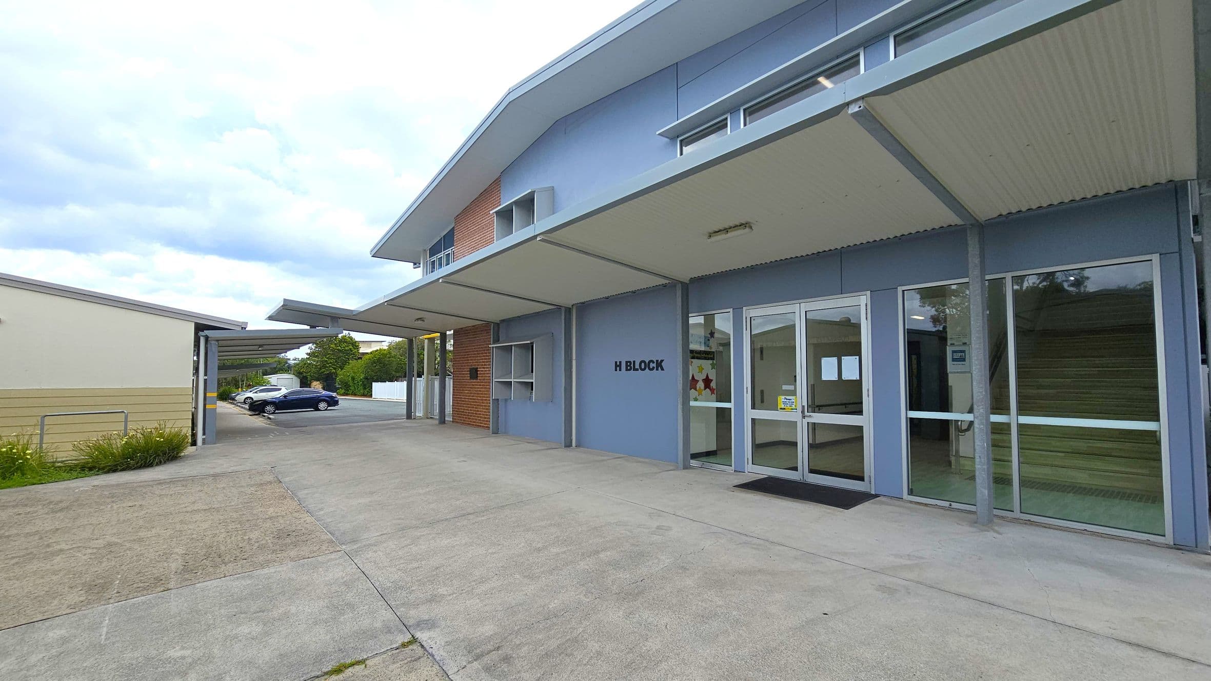 A modern building labelled "H Block" with a light blue exterior and large glass doors on the right. The building has a sloped roof with an overhang. To the left, there is a covered walkway leading to a parking area with two cars, one white and one black. The ground is paved with concrete, and there is a small patch of grass near the walkway. The background features trees and a white fence.