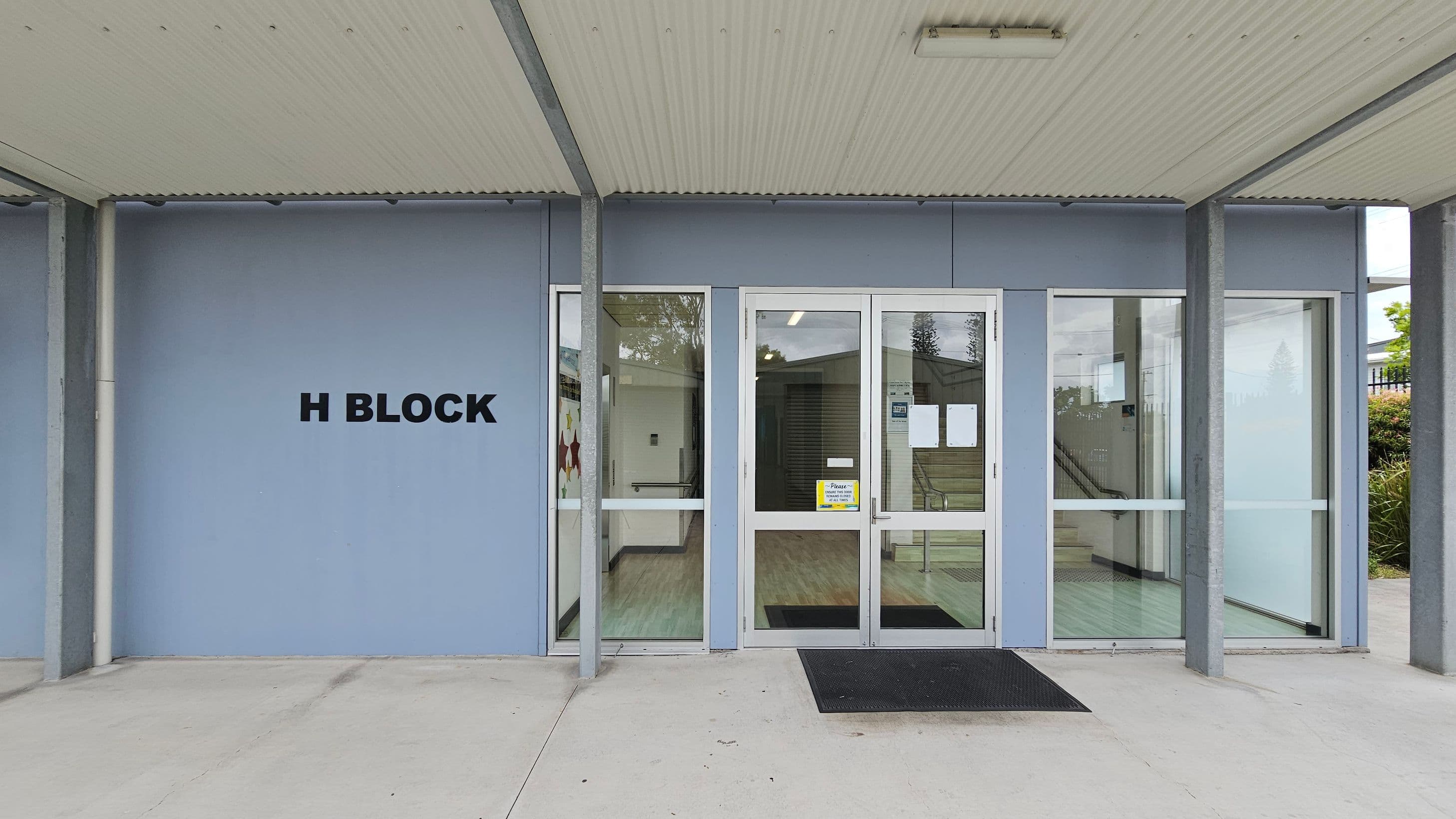 Entrance to a building labelled "H Block" on the left wall. The facade is light blue with large glass doors and windows. A black mat lies in front of the entrance. Inside, a corridor with a light-coloured floor is visible. The ceiling is corrugated and light-coloured, supported by metal beams. On the right, there are bushes outside the window.