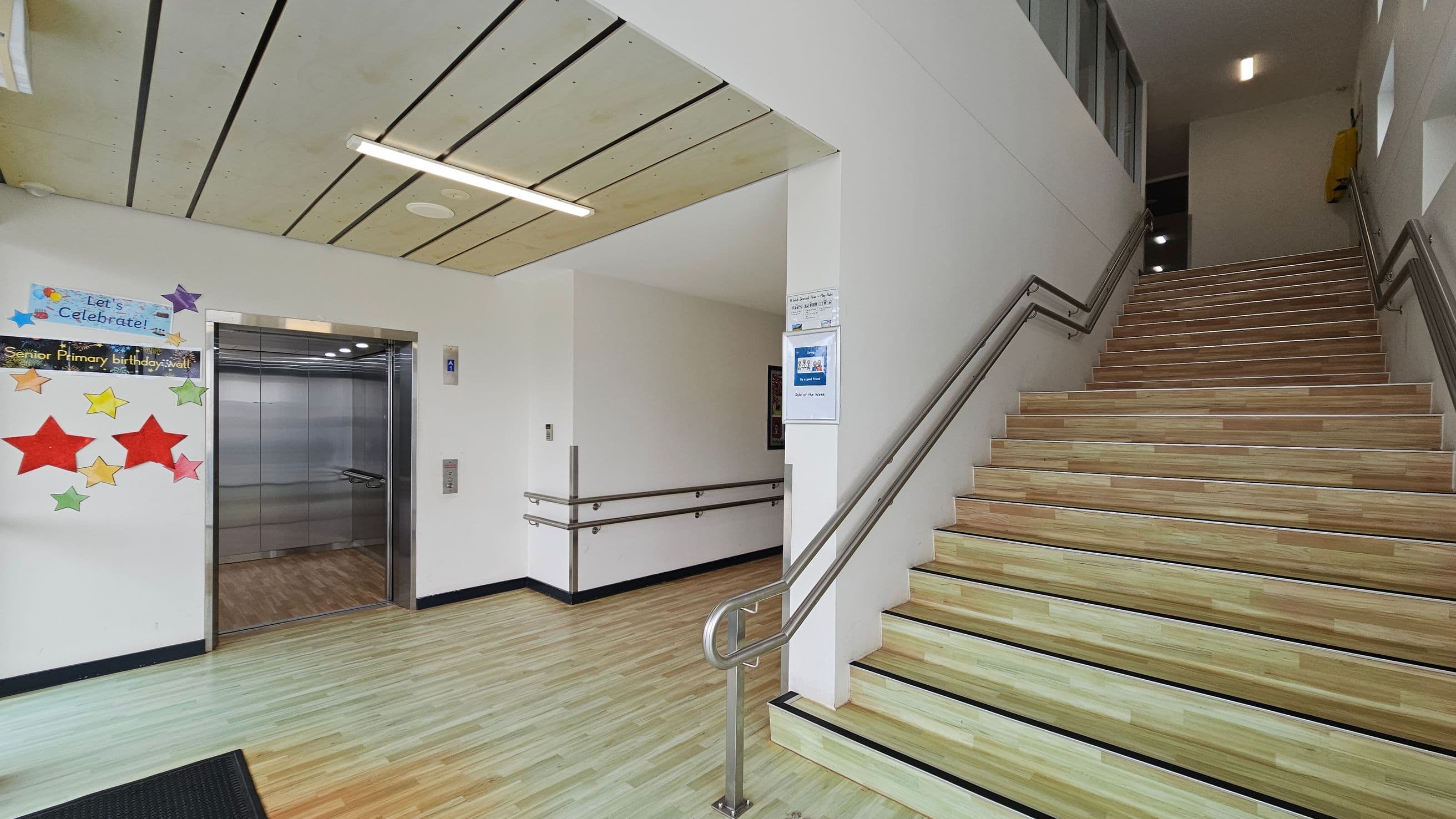 On the left, there is an open lift with a metallic interior and a colourful display of stars and a "Let's Celebrate!" sign on the wall. The floor is light wood with a black mat near the lift. On the right, a staircase with wooden steps and metal railings ascends. The ceiling is light-coloured with linear panels and a fluorescent light fixture. The walls are white, and there are informational posters near the stairs.
