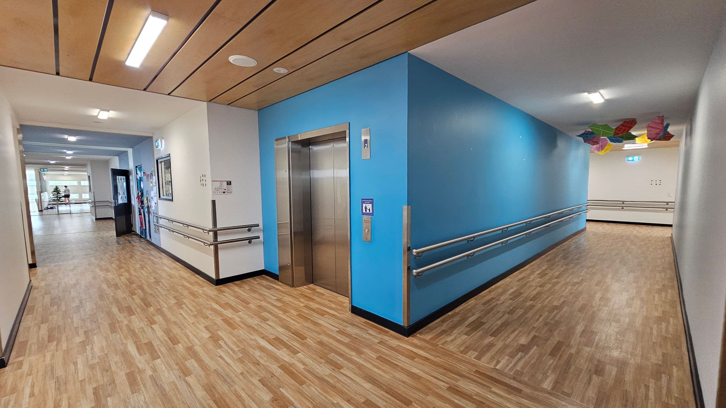 A corridor with wooden flooring and a wooden panelled ceiling. On the left, there's a lift with a blue wall and handrails. The corridor extends to the right, with colourful geometric shapes hanging from the ceiling. Walls are painted in light colours, and there are several overhead lights.