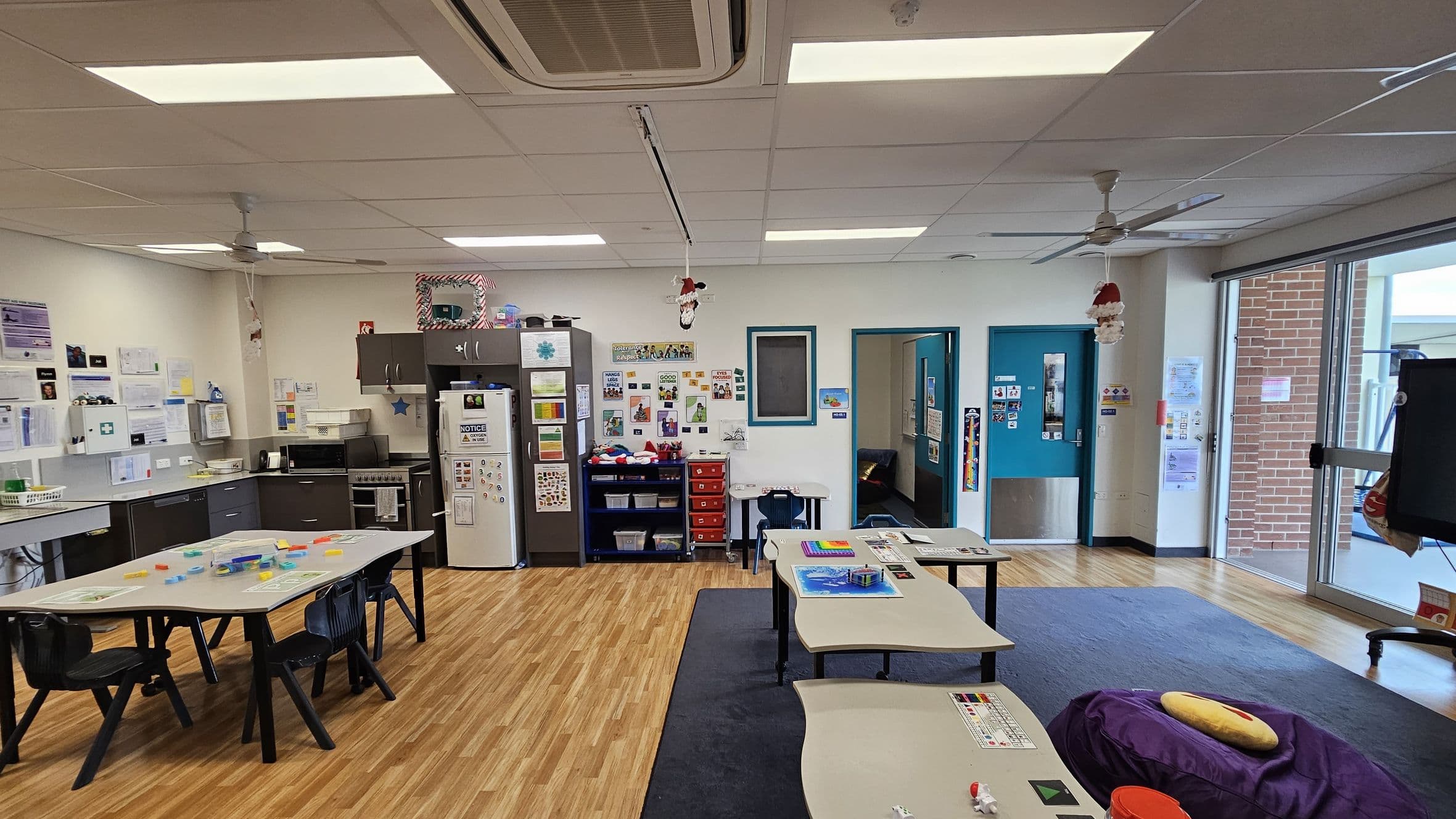 A classroom with a wooden floor and a white ceiling featuring fluorescent lights and ceiling fans. On the left, there is a kitchen area with a counter, sink, and appliances, including a fridge covered with notices. The back wall has educational posters and a small red drawer unit. In the centre, tables with chairs are arranged, some with educational materials. On the right, large windows and a door lead outside, with a brick wall visible through the glass.