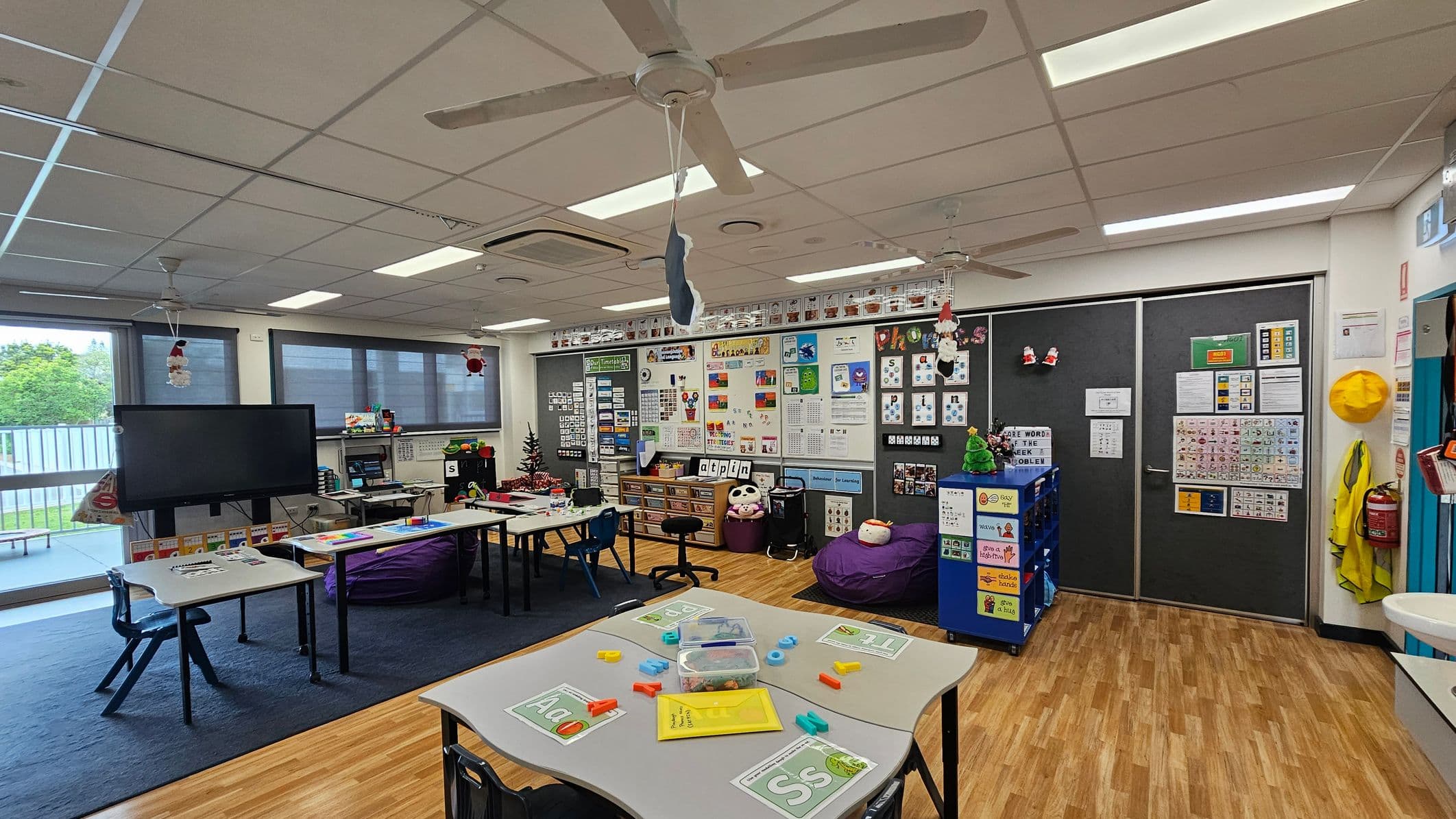A classroom with a wooden floor and a white ceiling featuring ceiling fans. On the left, there are tables with educational materials and a large screen near windows with blinds. The middle has a wall filled with educational posters and a small Christmas tree. On the right, there are dark sliding doors with more posters, a yellow hat, and a fire extinguisher. A blue storage unit and bean bags are also visible.
