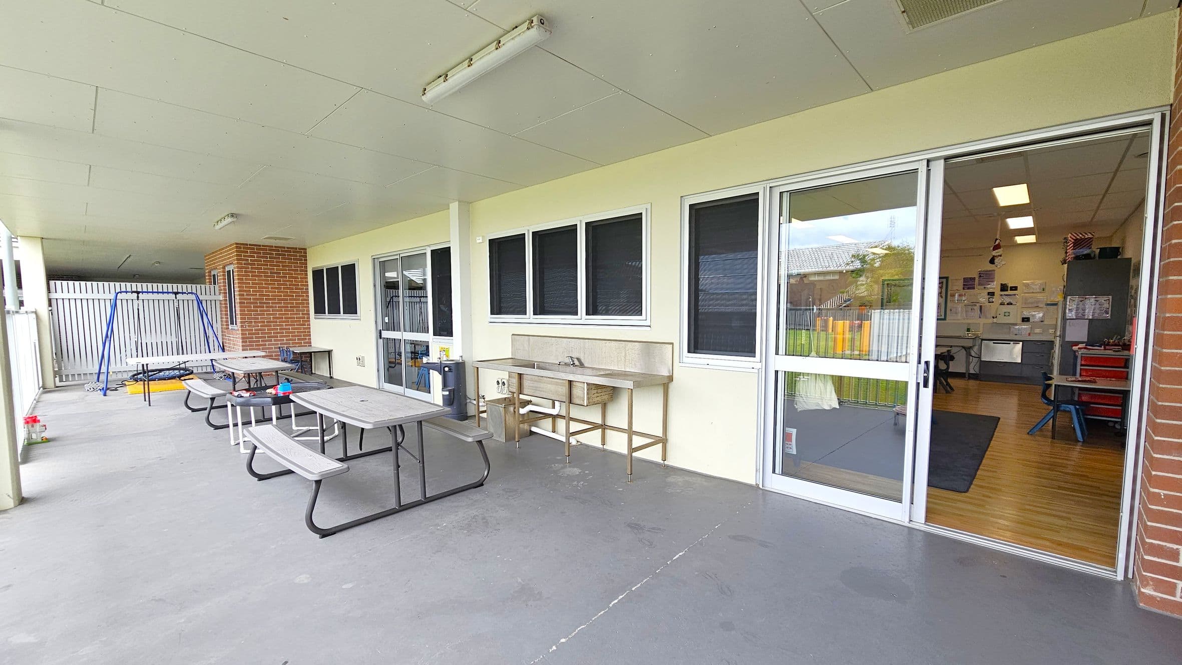 A covered outdoor area with a concrete floor and a ceiling with fluorescent lights. On the left, there is a blue swing set and several picnic tables with benches. In the middle, a stainless steel sink is mounted against the wall. On the right, large sliding glass doors lead into an indoor room with wooden flooring, visible tables, chairs, and a cabinet. The walls are a light colour, and there is a brick column near the entrance.
