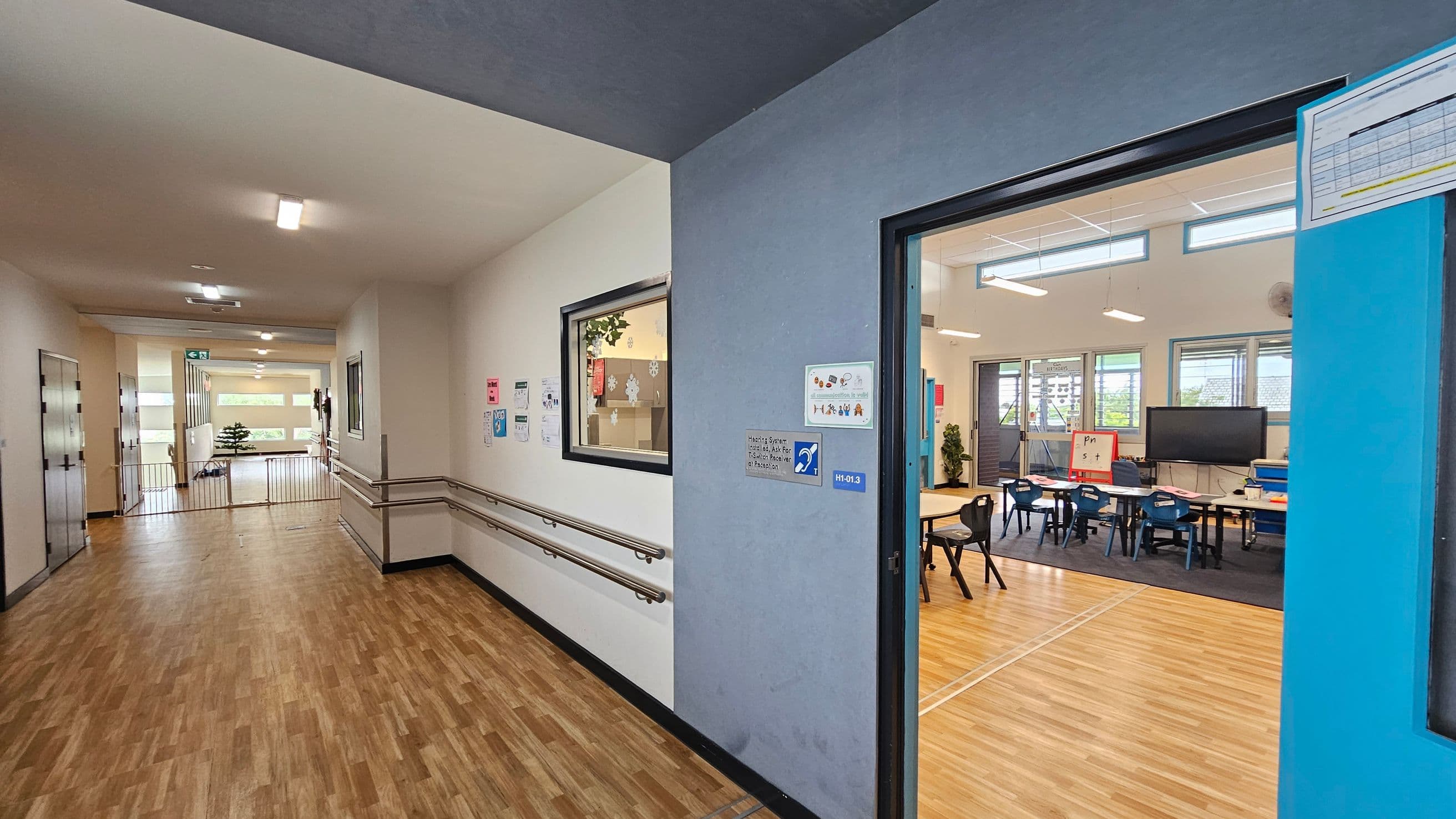 A school corridor with wooden flooring and white walls. On the left, there are handrails along the wall and a window with a view into a room. The corridor leads to a bright area with large windows and a small plant. On the right, an open door reveals a classroom with tables, chairs, a large screen, and a whiteboard. The classroom has light wooden flooring and several windows allowing natural light. Ceiling lights are visible in both the corridor and the