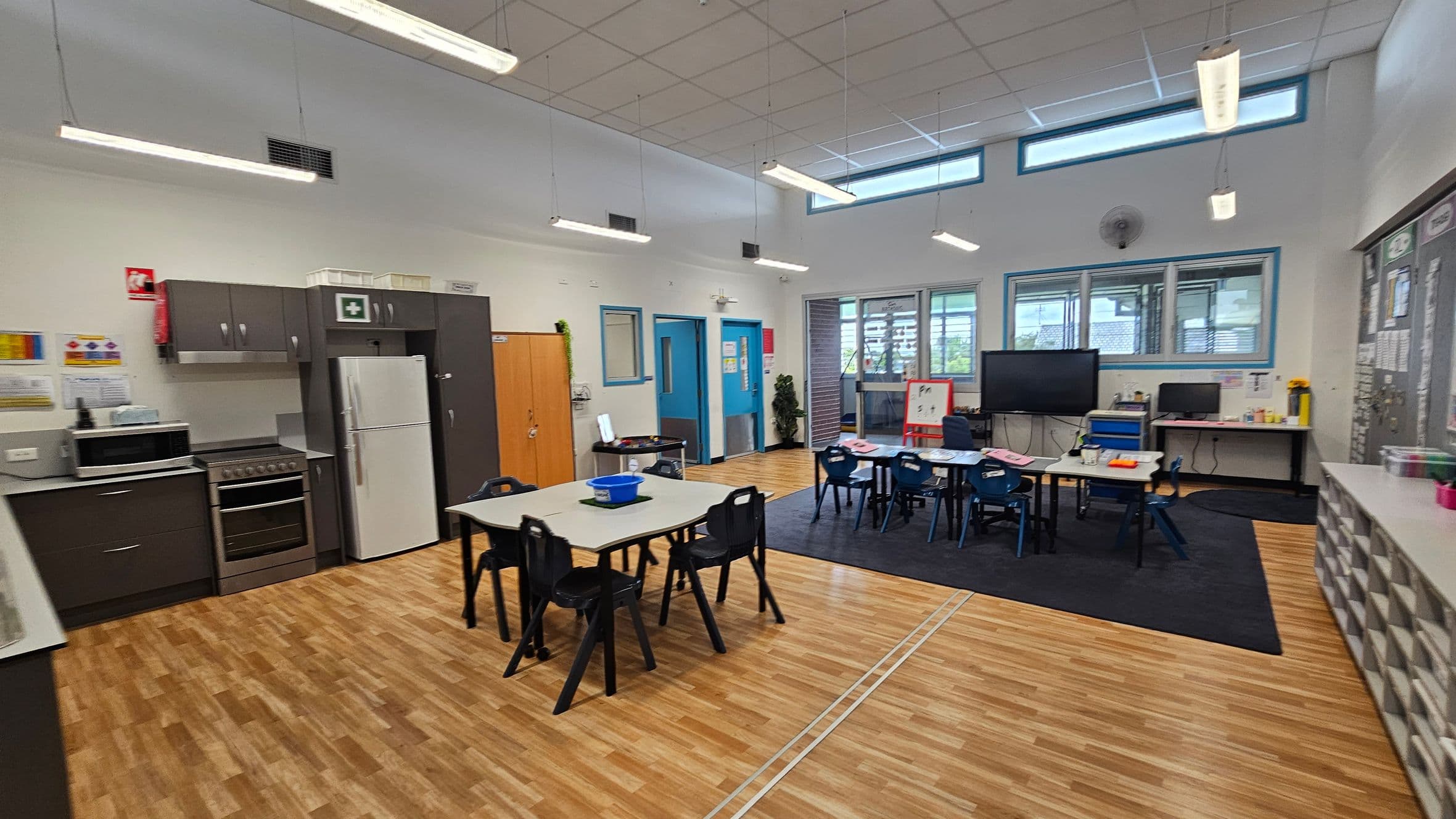 A classroom with a wooden floor and a white ceiling featuring fluorescent lights. On the left, there is a kitchen area with a stove, microwave, and refrigerator. In the centre, tables and chairs are arranged for students. The right side has a large screen, a computer on a desk, and a whiteboard. Windows line the upper part of the walls, allowing natural light. Storage units with multiple compartments are visible on the far right.