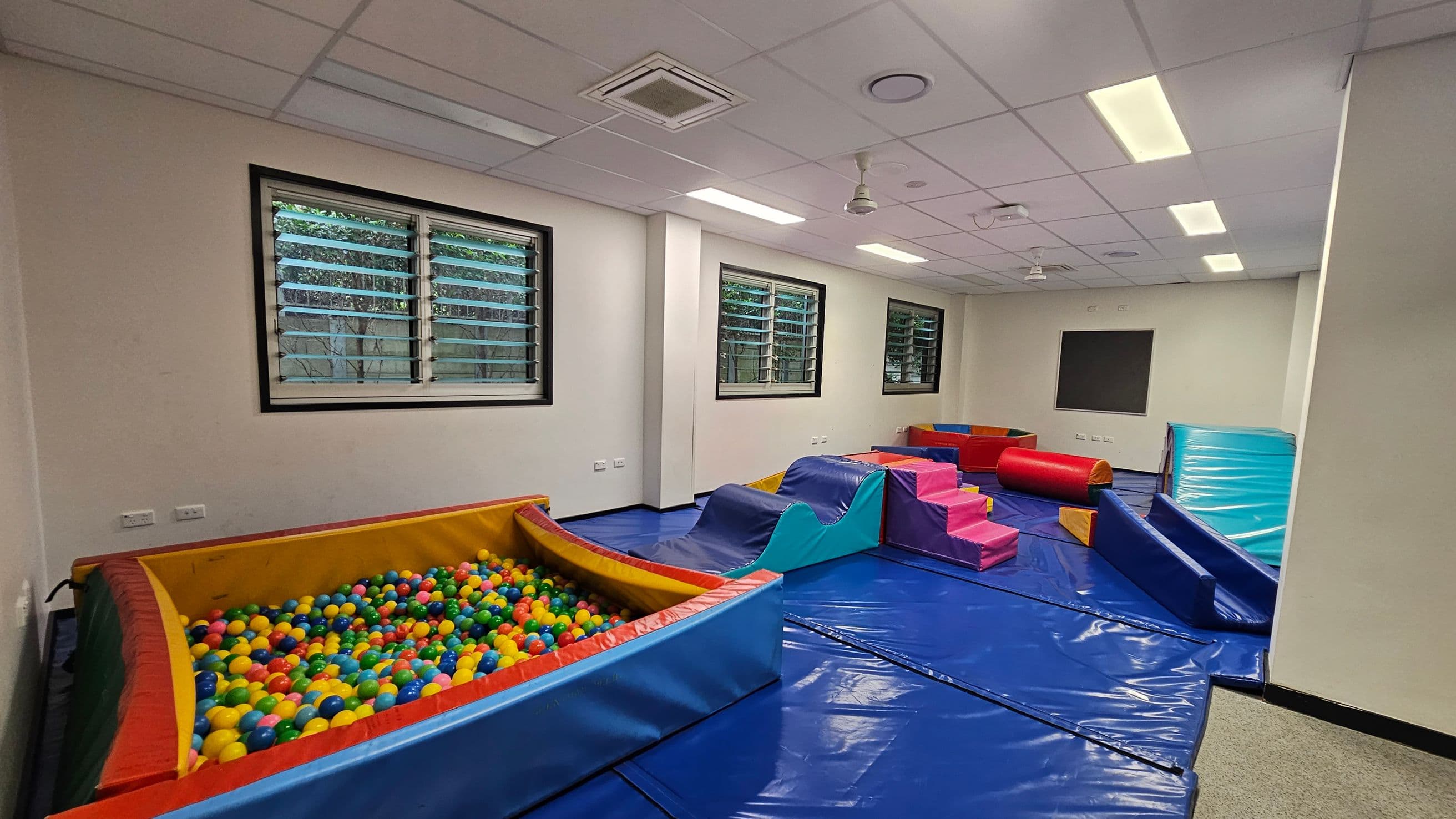 A playroom with a ball pit filled with colourful balls on the left. In the centre, there are soft play structures including a blue wave-shaped ramp and pink steps. On the right, a red cylindrical cushion and a blue mat are visible. The floor is covered with blue mats, and the ceiling has white panels with fluorescent lights. Three windows with horizontal blinds are on the back wall, allowing natural light to enter.