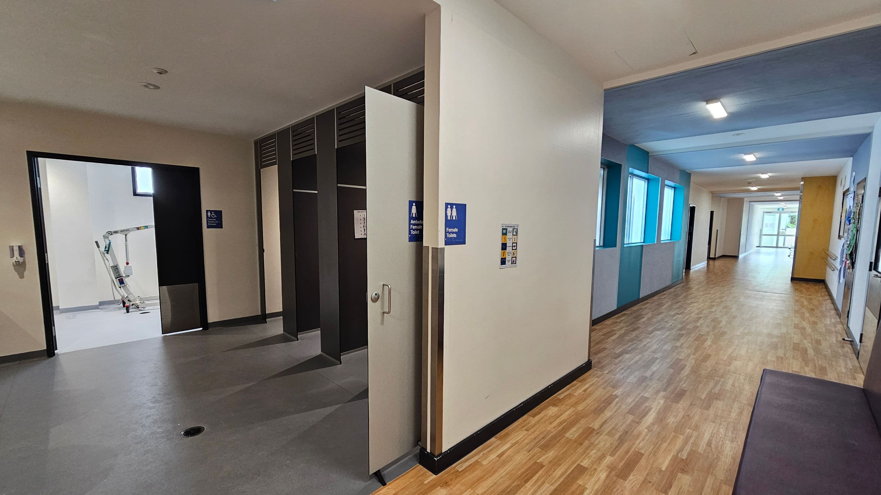 On the left, there is an open door leading to a room with a grey floor and a piece of equipment visible inside. The door has signs indicating accessible and female toilets. On the right, a corridor with a wooden floor extends into the distance, featuring several windows with blue frames on the left wall. The ceiling is white with recessed lighting. The corridor leads to a glass door at the far end.