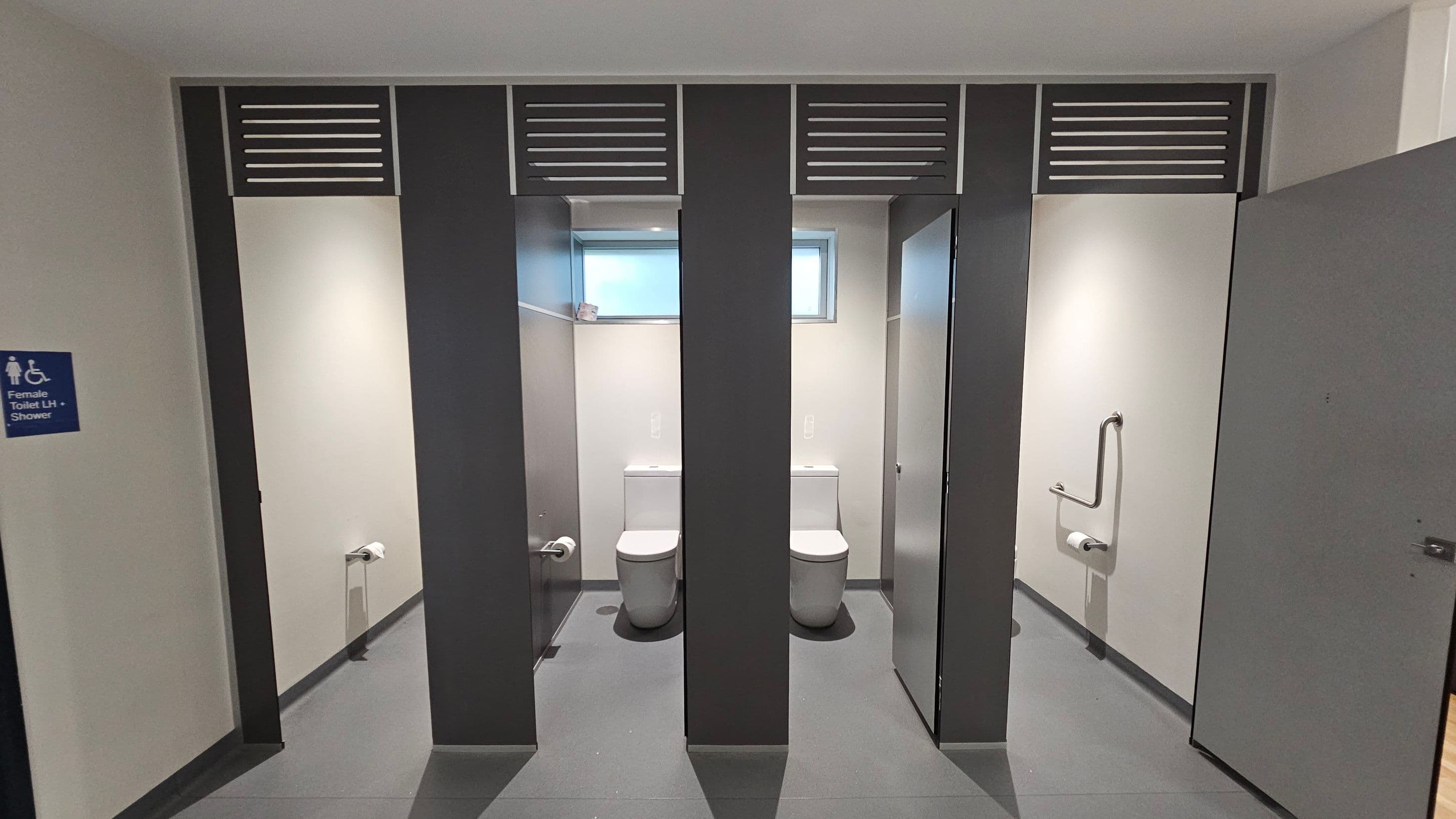 A public restroom with two toilet cubicles in the centre, each with a white toilet and a small window above. The cubicles have grey partitions with ventilation panels at the top. On the right, a door is partially open, revealing a grab bar and toilet paper holder. On the left, a sign indicates a female toilet with shower facilities. The floor is grey, and the walls are light-coloured.