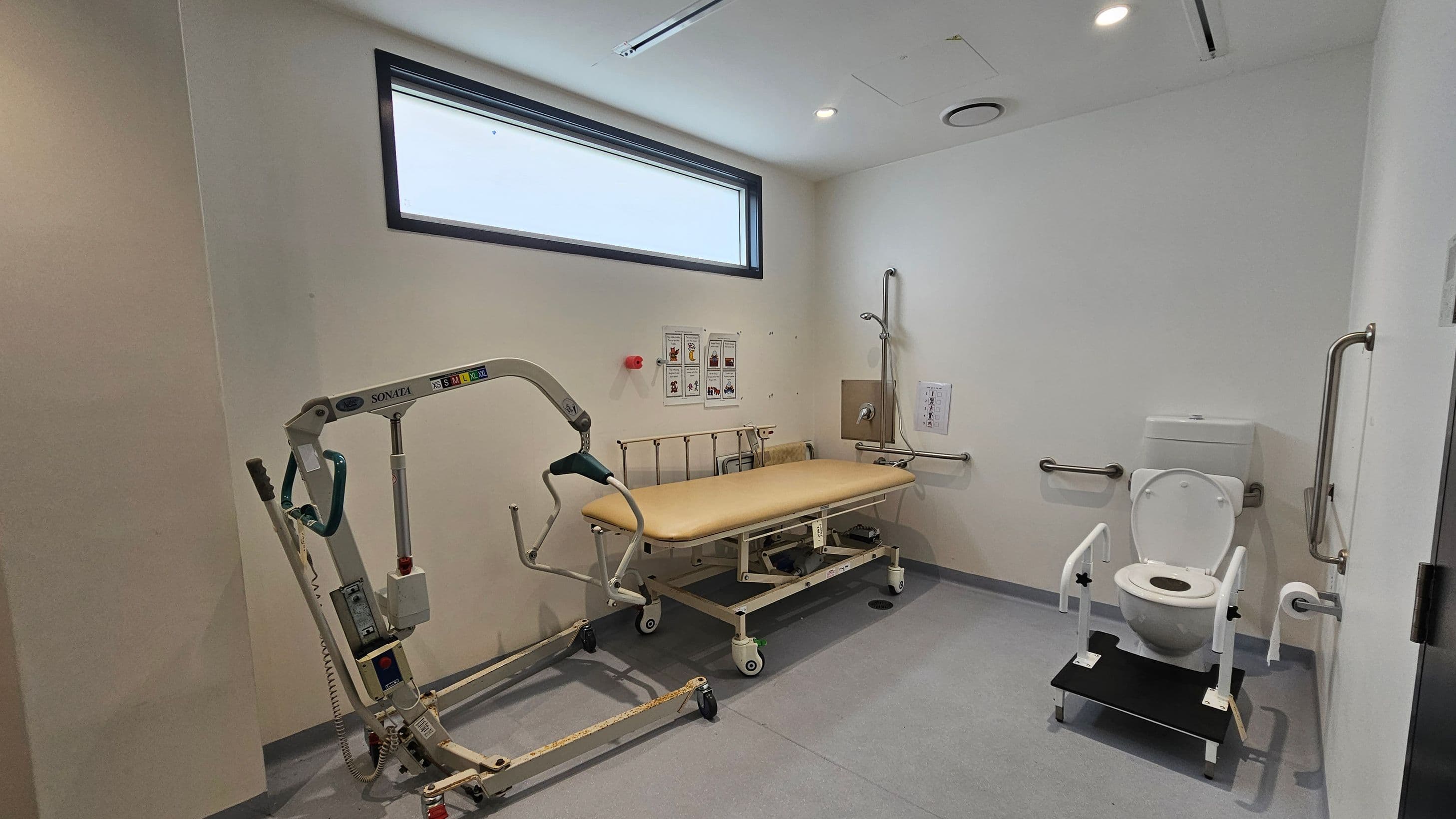 A room with a grey floor and white walls. On the left, there is a mobile hoist with a green handle. In the centre, a beige adjustable bed with side rails is positioned against the wall, beneath a rectangular window. On the right, a toilet with support bars and a foot platform is installed. The walls have several grab bars and a shower head. The ceiling is white with recessed lighting and ventilation.