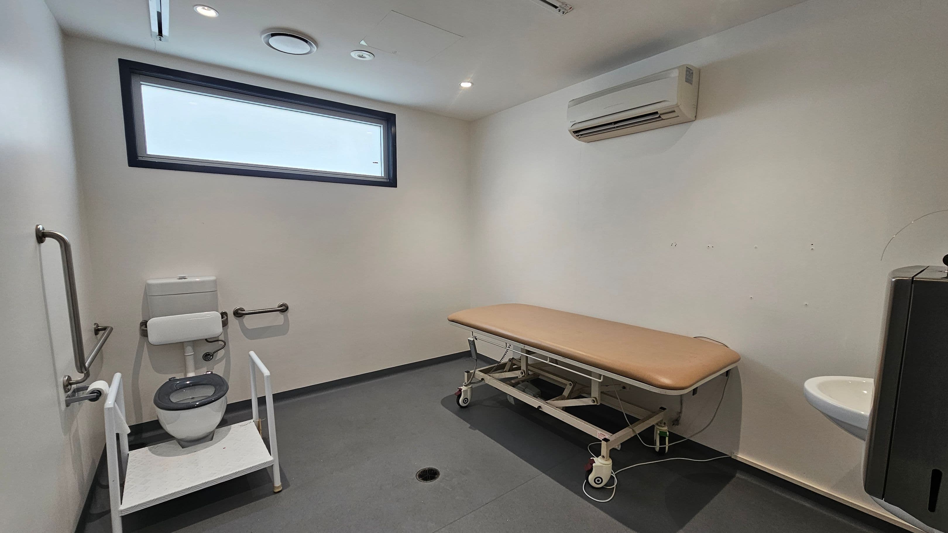 A room with a grey floor and white walls. On the left, there's a toilet with support rails and a white frame. In the middle, a brown cushioned examination table on wheels. On the right, a small white sink with a black hand dryer above it. A rectangular window is high on the left wall, and an air conditioning unit is mounted on the right wall. The ceiling has recessed lighting and ventilation fixtures.