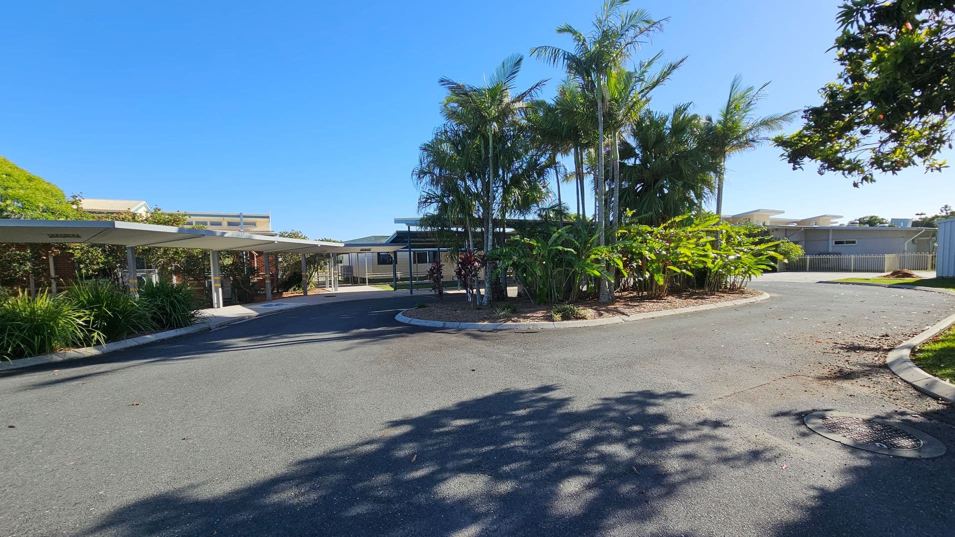 A paved driveway curves around a central island filled with tall palm trees and lush green plants. On the left, there are covered parking spaces with metal roofs, bordered by more greenery. In the background, a low building with a flat roof is visible, partially obscured by the trees. The area is well-maintained, with shadows of trees cast on the pavement.