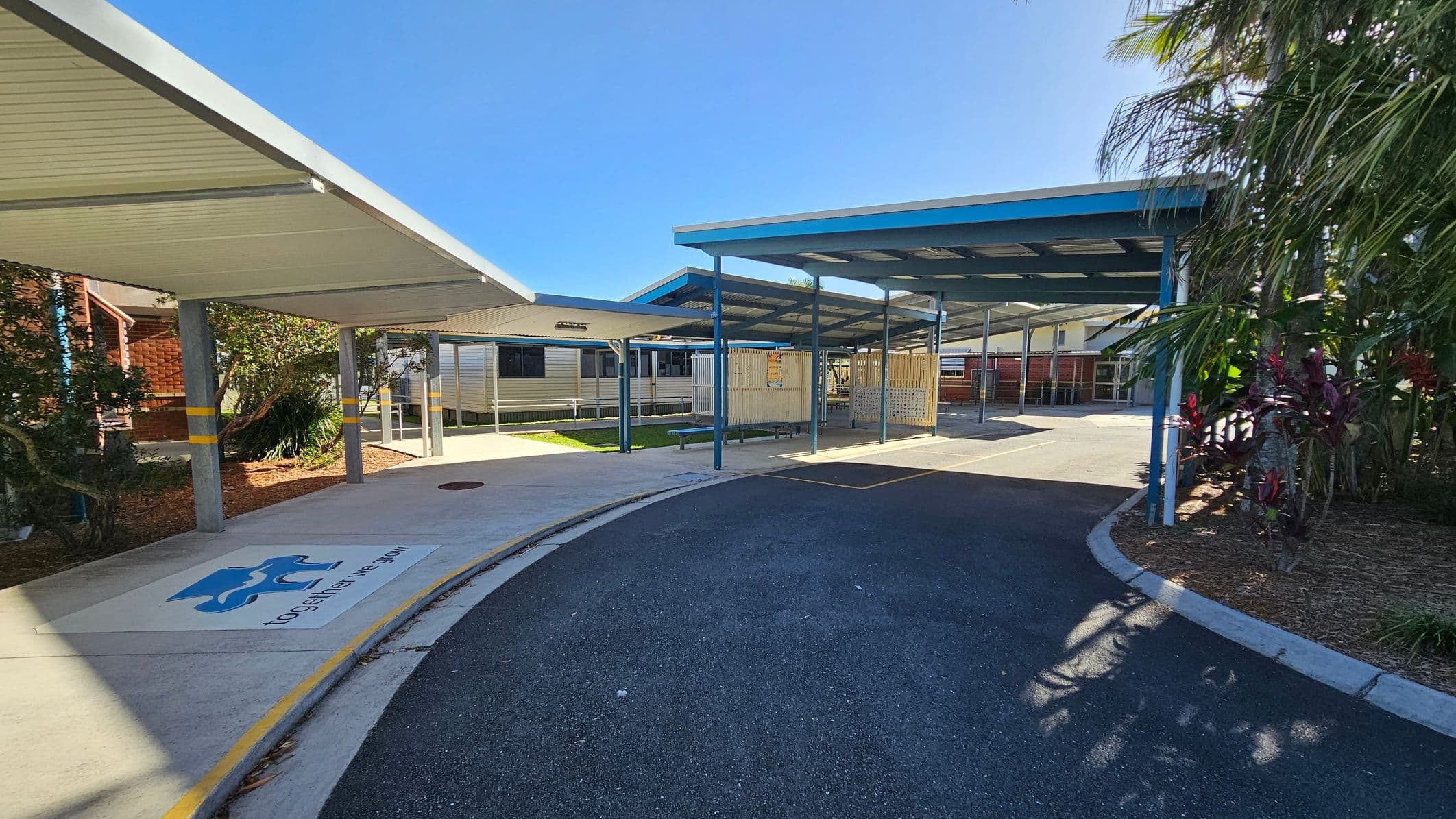 Covered walkways with blue and white roofs extend from the left to the centre, supported by grey pillars. A pathway curves from the foreground to the centre, with a logo and text on the ground. On the right, there are plants and trees. In the background, there are buildings with metal siding and windows. The ground is a mix of concrete and asphalt.