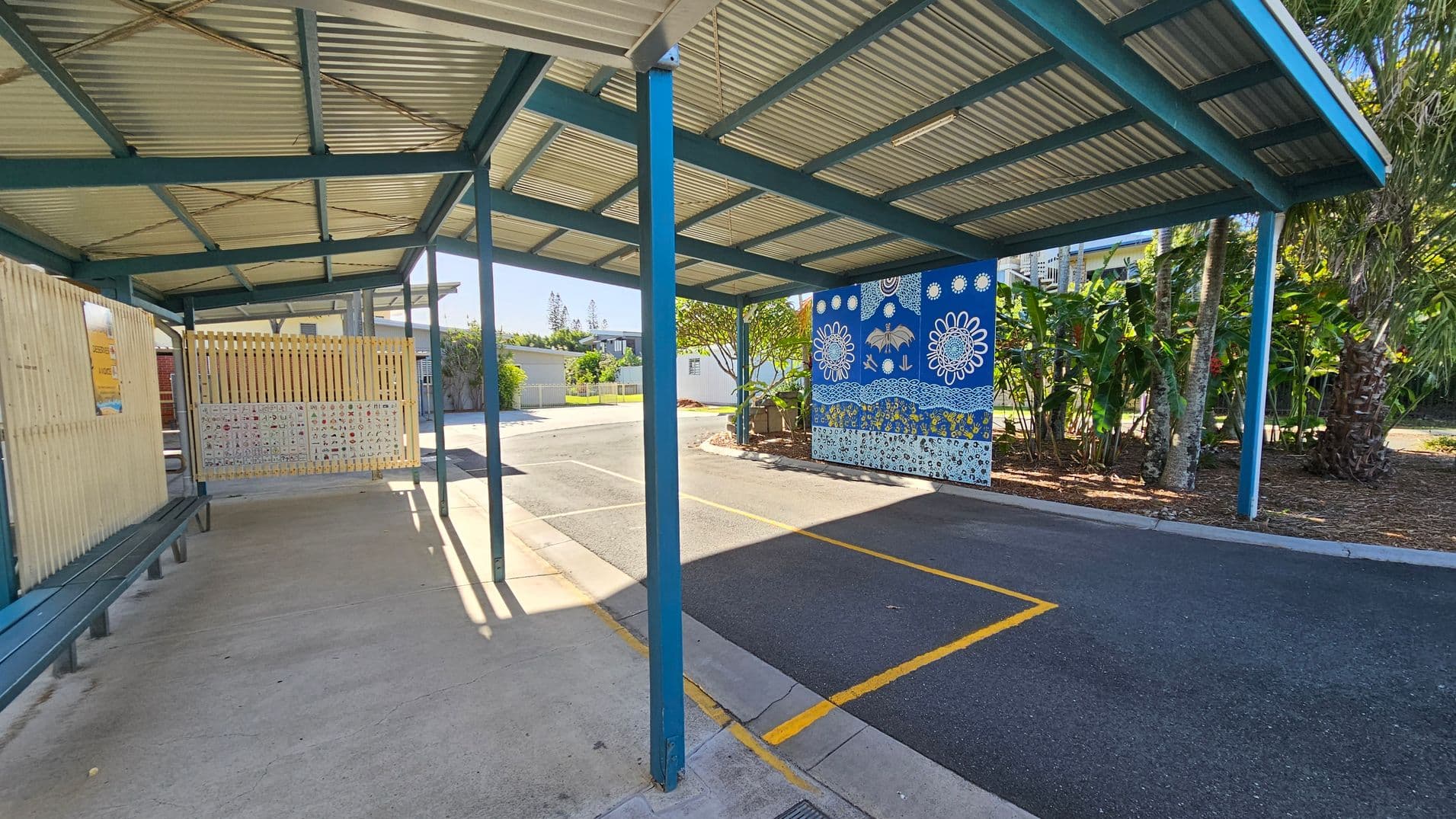 A covered walkway with a corrugated metal roof supported by blue beams. On the left, there is a wooden fence with a bench beneath it. The ground is a mix of concrete and asphalt. On the right, a vibrant blue mural with white floral and abstract designs is visible. Behind the mural, there are lush green plants and trees. The area is well-lit with sunlight casting shadows on the ground.