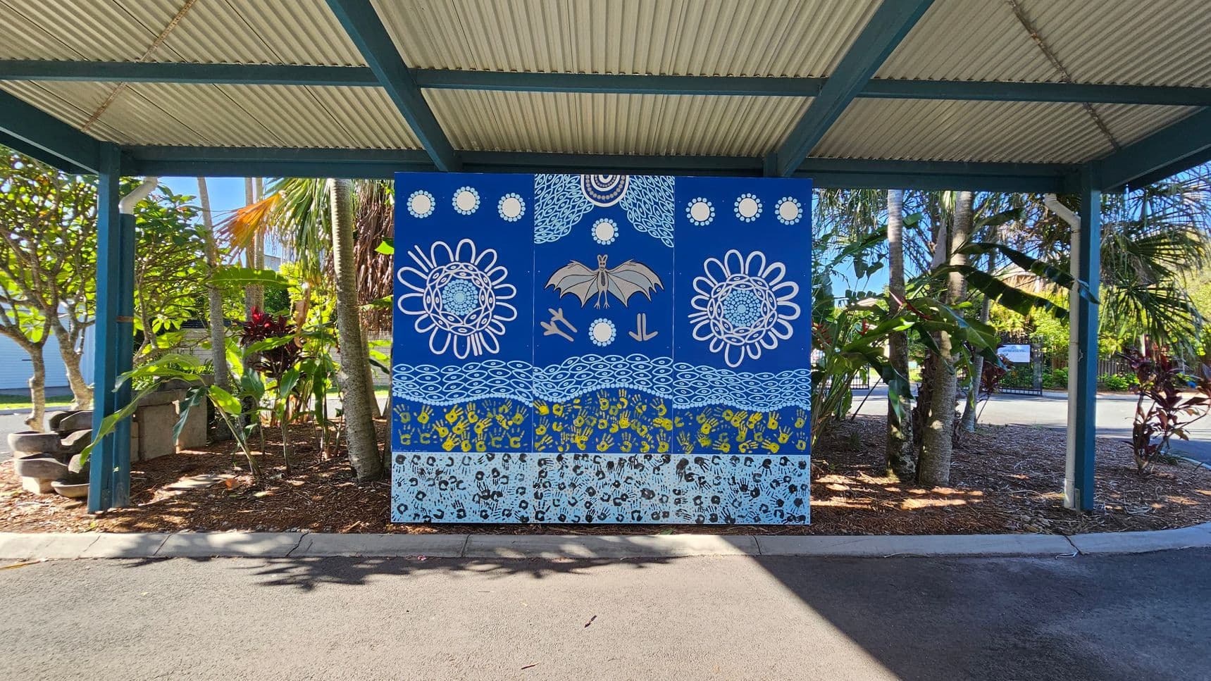 A vibrant mural is centred under a corrugated metal roof supported by green beams. The mural features a blue background with intricate designs, including a bat, circular patterns, and handprints in yellow and black. Surrounding the mural are lush tropical plants with broad leaves and some red foliage. The ground is covered with mulch and the foreground has a paved area.