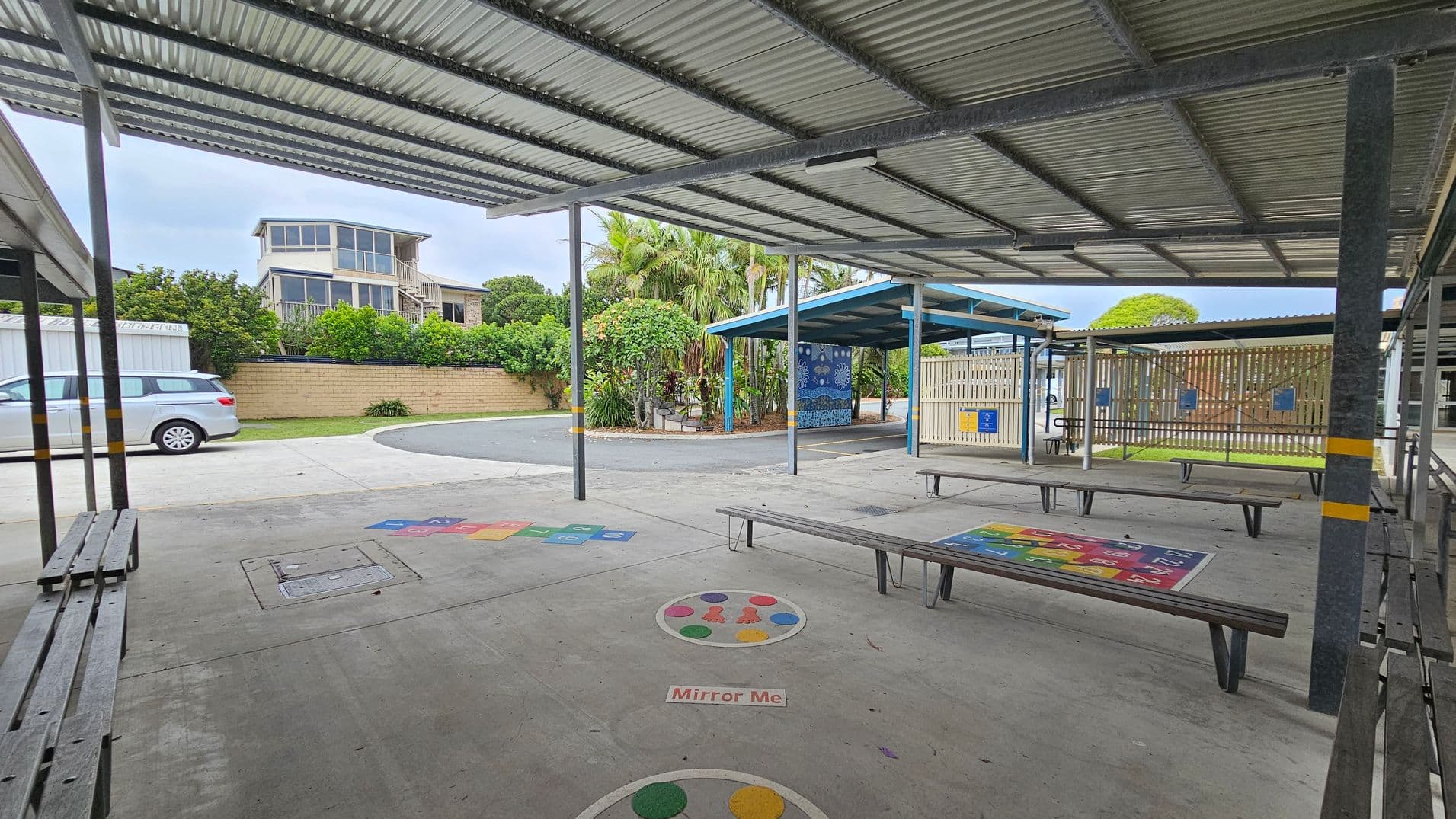 A covered outdoor area with metal roofing and benches. The concrete floor has colourful game markings, including a "Mirror Me" circle. In the background, there's a two-storey building with large windows, a parked silver car, and a brick wall with lush greenery. To the right, a fenced area with a blue-roofed shelter and more benches. Palm trees and other plants are visible near the centre.