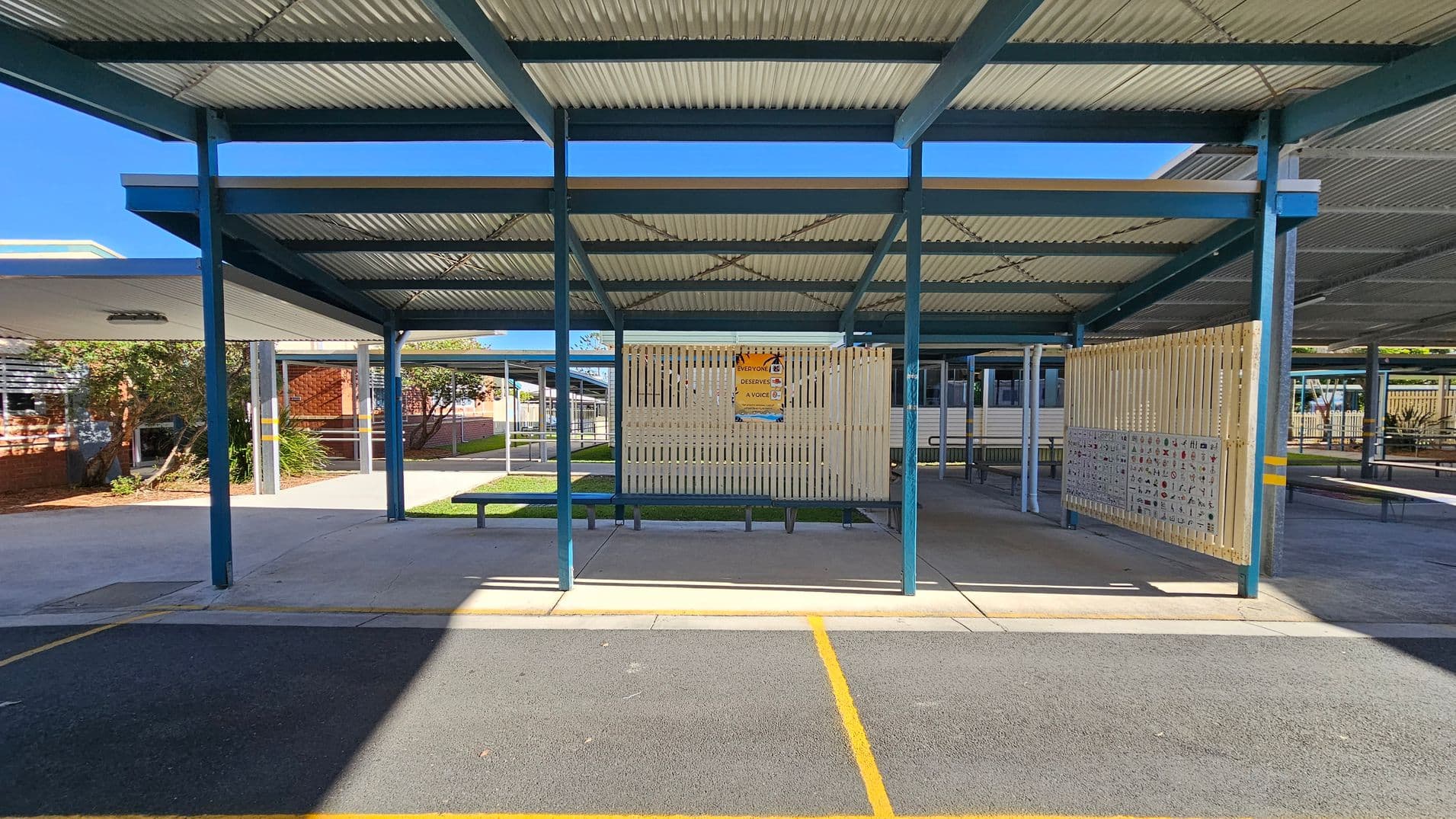 A covered outdoor area with a corrugated metal roof supported by blue metal beams. In the centre, a wooden slatted partition displays a poster. The ground is a mix of concrete and a textured surface with yellow lines. To the left, there are plants and a brick building with windows. The right side shows more covered walkways and benches. The background includes additional structures and greenery.