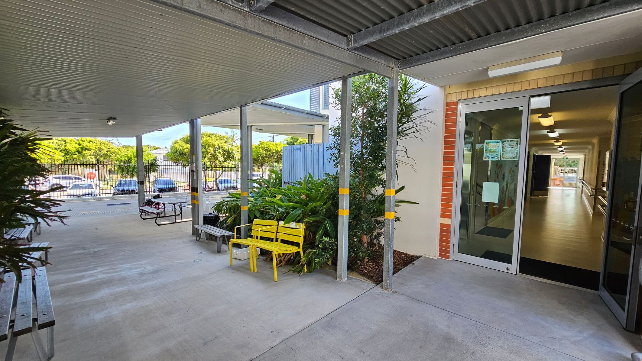 A covered outdoor area with a concrete floor and a corrugated metal ceiling. On the right, there is a glass door leading to a hallway with wooden flooring and ceiling lights. Near the door, there are green plants and a yellow bench. On the left, there are metal benches and a picnic table. In the background, a fence and parked cars are visible, along with trees.
