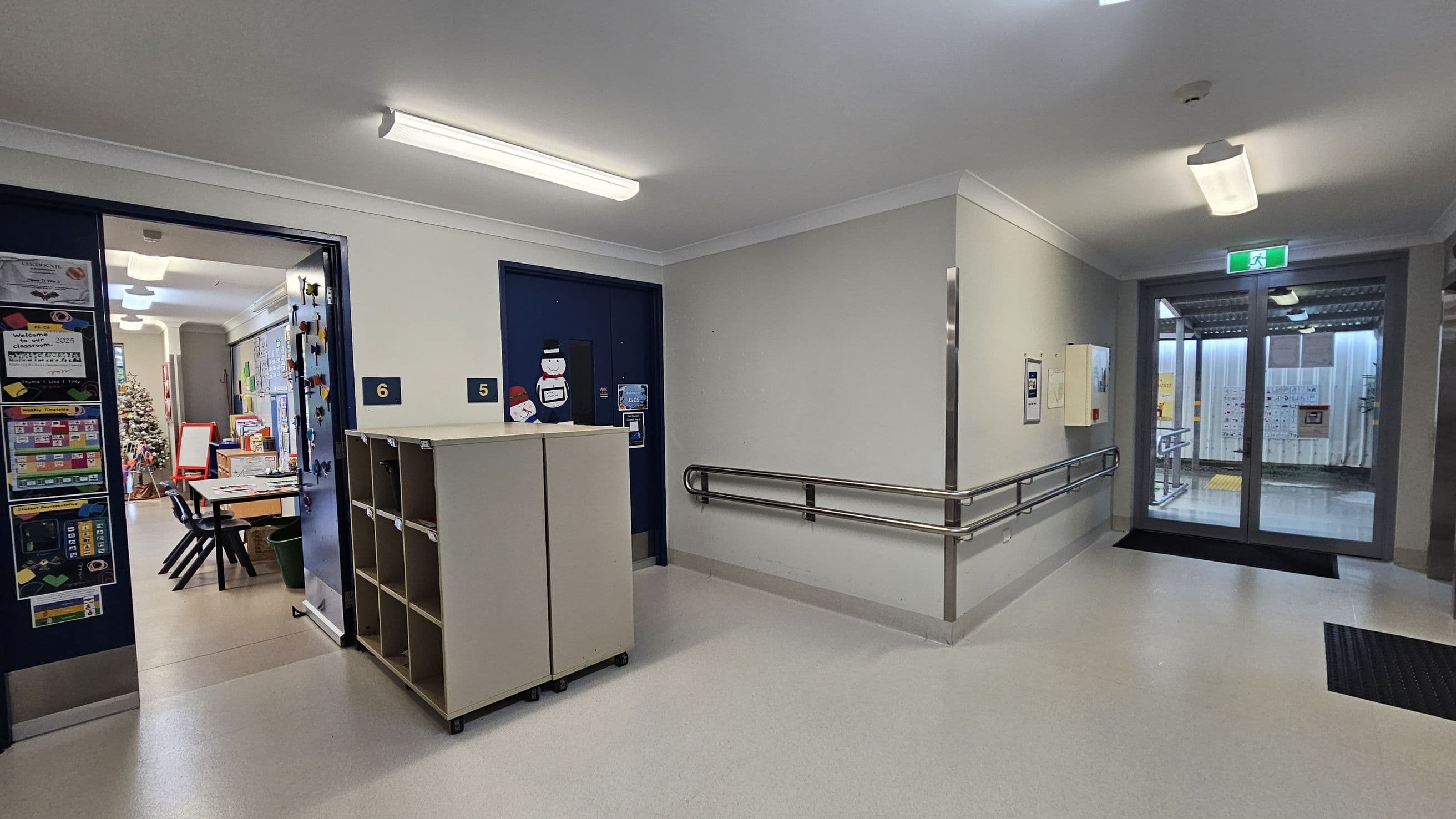 A hallway with a light grey floor and white ceiling featuring fluorescent lights. On the left, an open door leads to a classroom with tables, chairs, and a Christmas tree. The door is marked with the number 6. Adjacent is a closed door with the number 5, decorated with snowman cut-outs. A small shelving unit is positioned between the doors. On the right, a handrail runs along the wall leading to a glass door with an exit sign above, opening to an outdoor area.