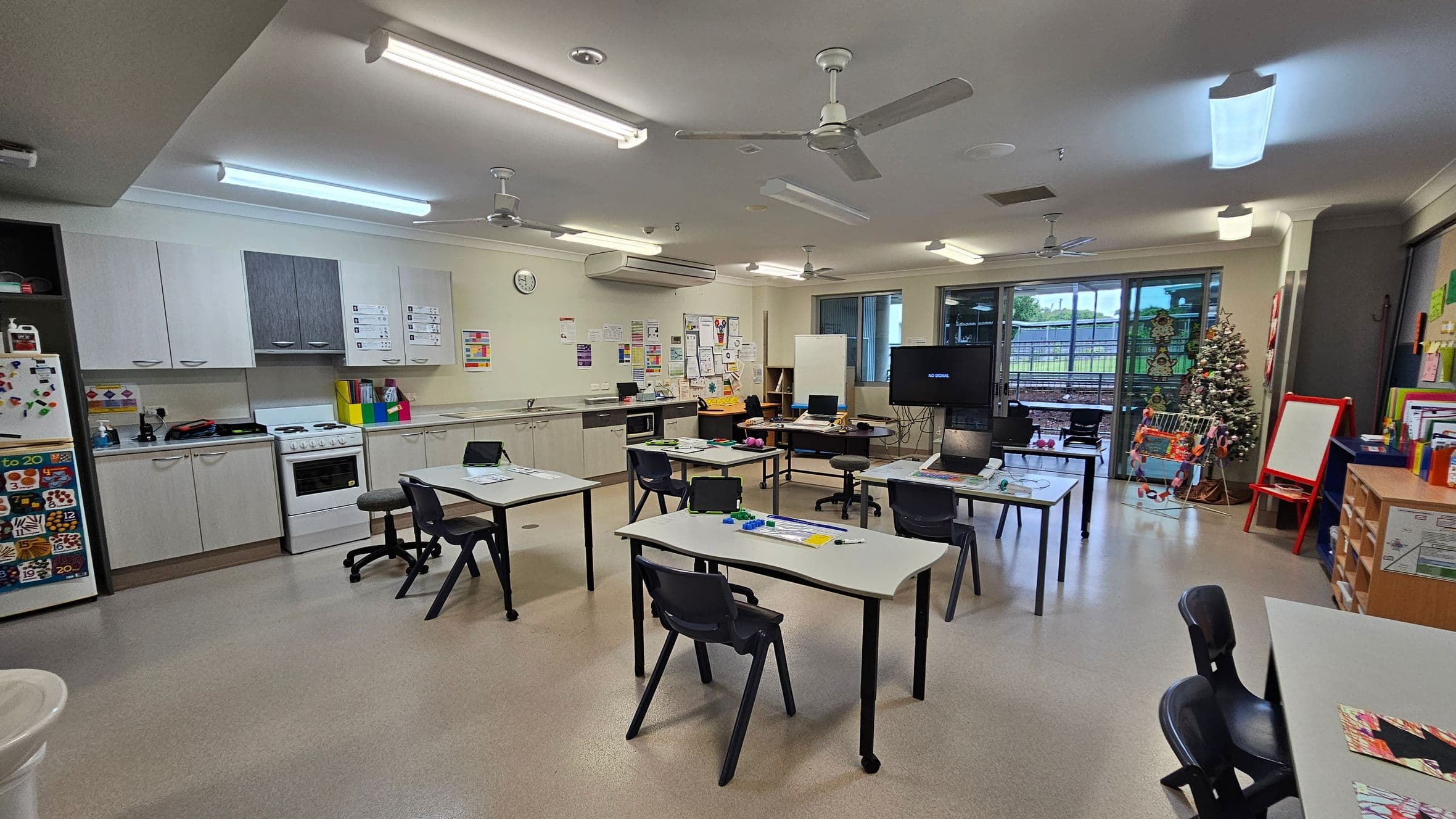 A classroom with a kitchen area on the left, featuring white cabinets, a stove, and a fridge adorned with colourful magnets. In the centre, several tables and chairs are arranged, some with laptops and educational materials. The right side has a Christmas tree and a red easel. The back wall has large windows and a door leading outside. The ceiling has fluorescent lights and ceiling fans, and the floor is a light-coloured linoleum.