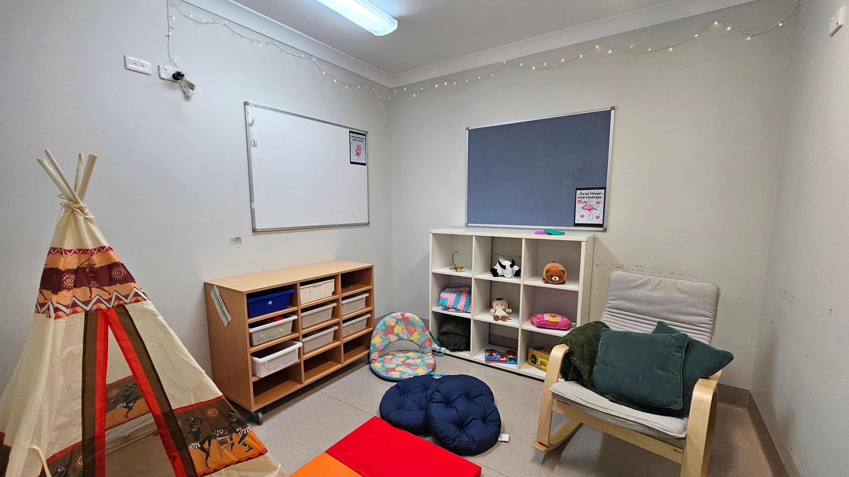 A small room with a teepee on the left, decorated with patterns in brown, orange, and red. In the centre, a wooden shelf unit with various storage boxes. To the right, a white cubby shelf holds toys and books. A cushioned chair with green pillows is nearby. The walls have a whiteboard and a blue notice board. The ceiling has a fluorescent light and string lights along the edges. The floor is carpeted in a neutral tone.