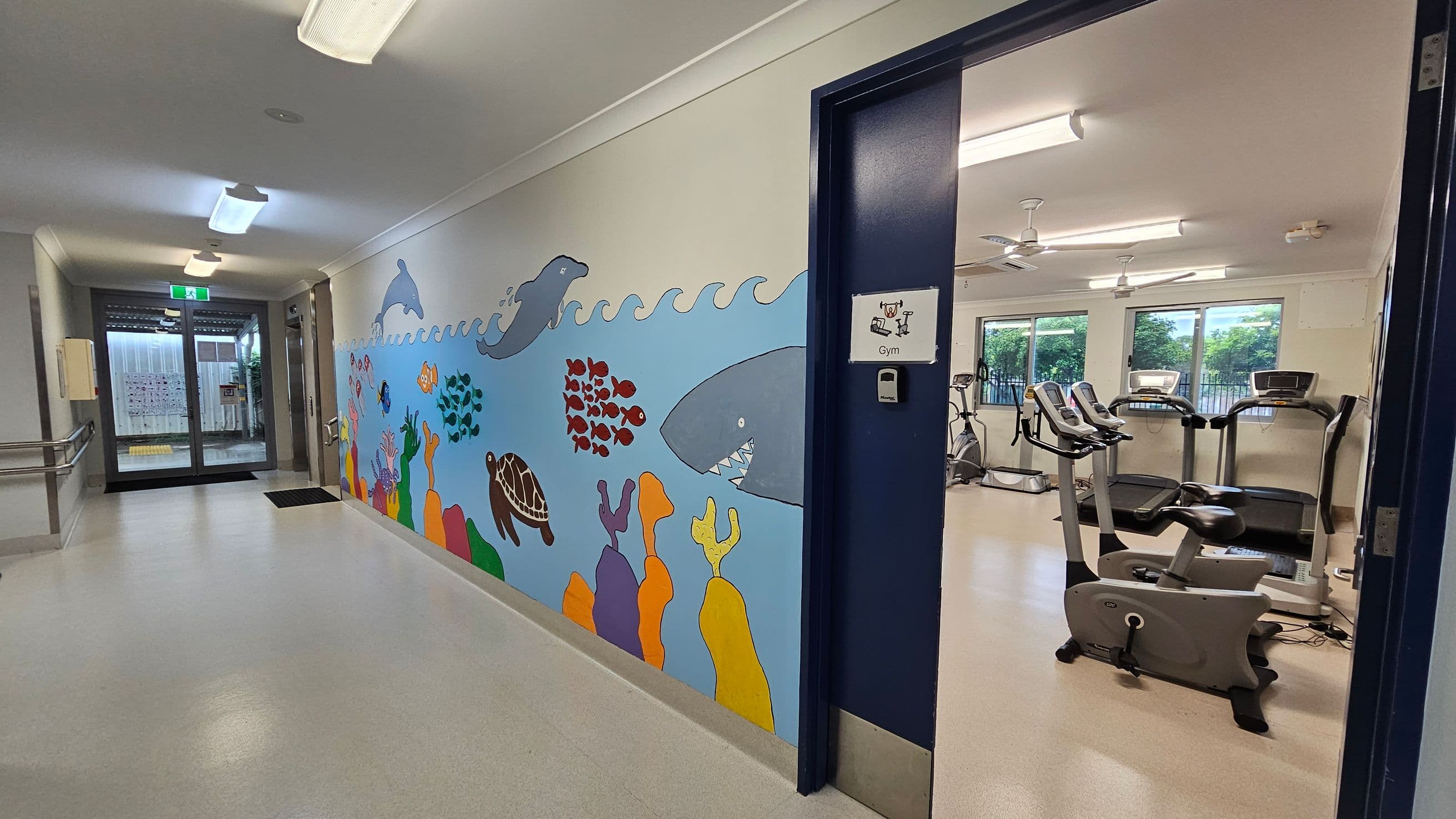 A hallway with a light-coloured floor and ceiling, featuring a colourful mural on the left wall depicting sea creatures like dolphins, fish, a turtle, and a shark. On the right, an open door leads to a gym with exercise equipment, including treadmills and stationary bikes. The gym has large windows with a view of greenery outside. The hallway ends with glass doors leading outside, and there are ceiling lights and a handrail on the left.