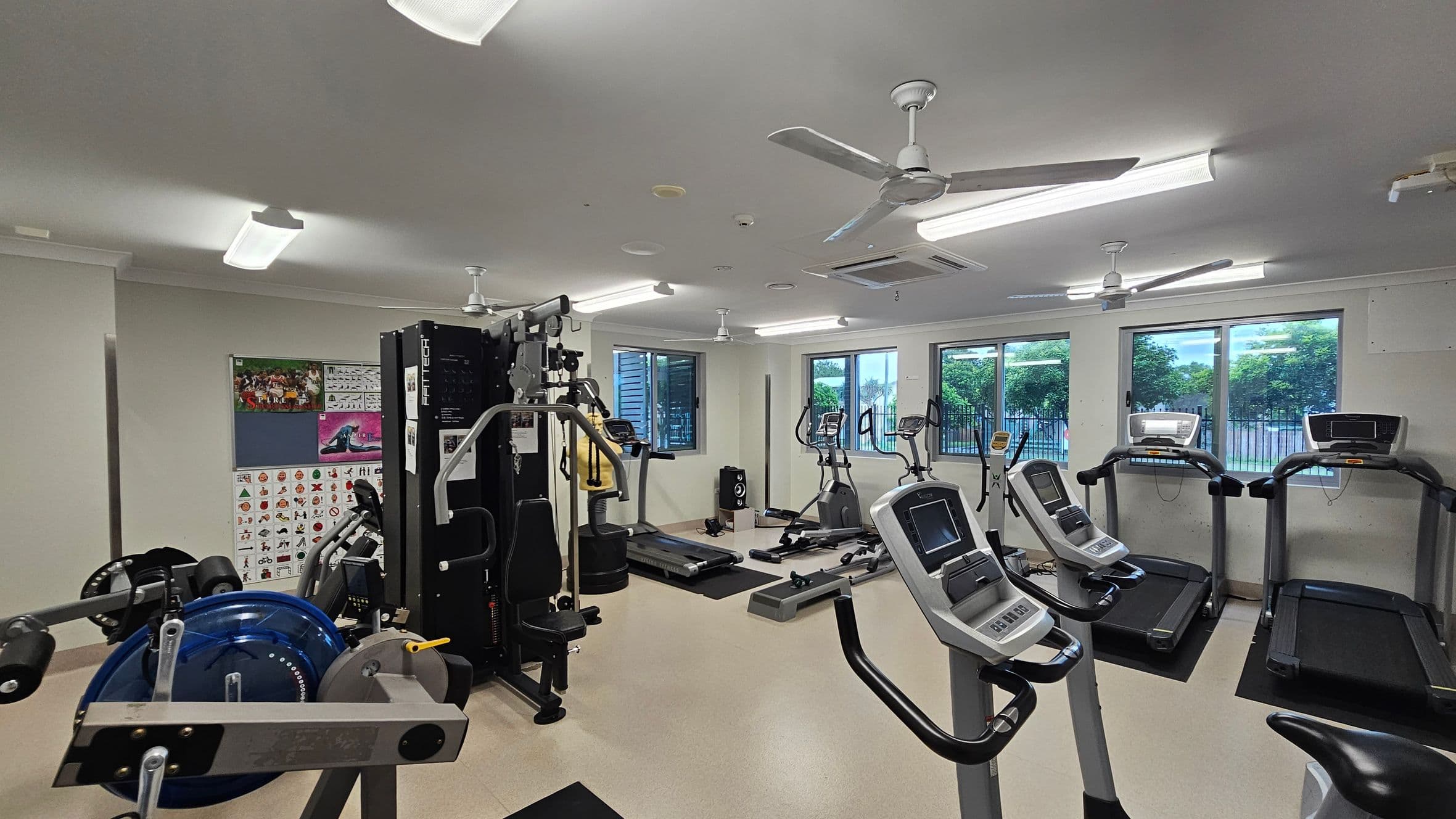 A gym with various exercise equipment. On the left, there is a multi-gym machine and weightlifting equipment. In the middle, there are several exercise bikes and a treadmill. On the right, there are more treadmills. The walls have posters and windows showing greenery outside. The ceiling has fluorescent lights and ceiling fans. The floor is light-coloured and smooth.