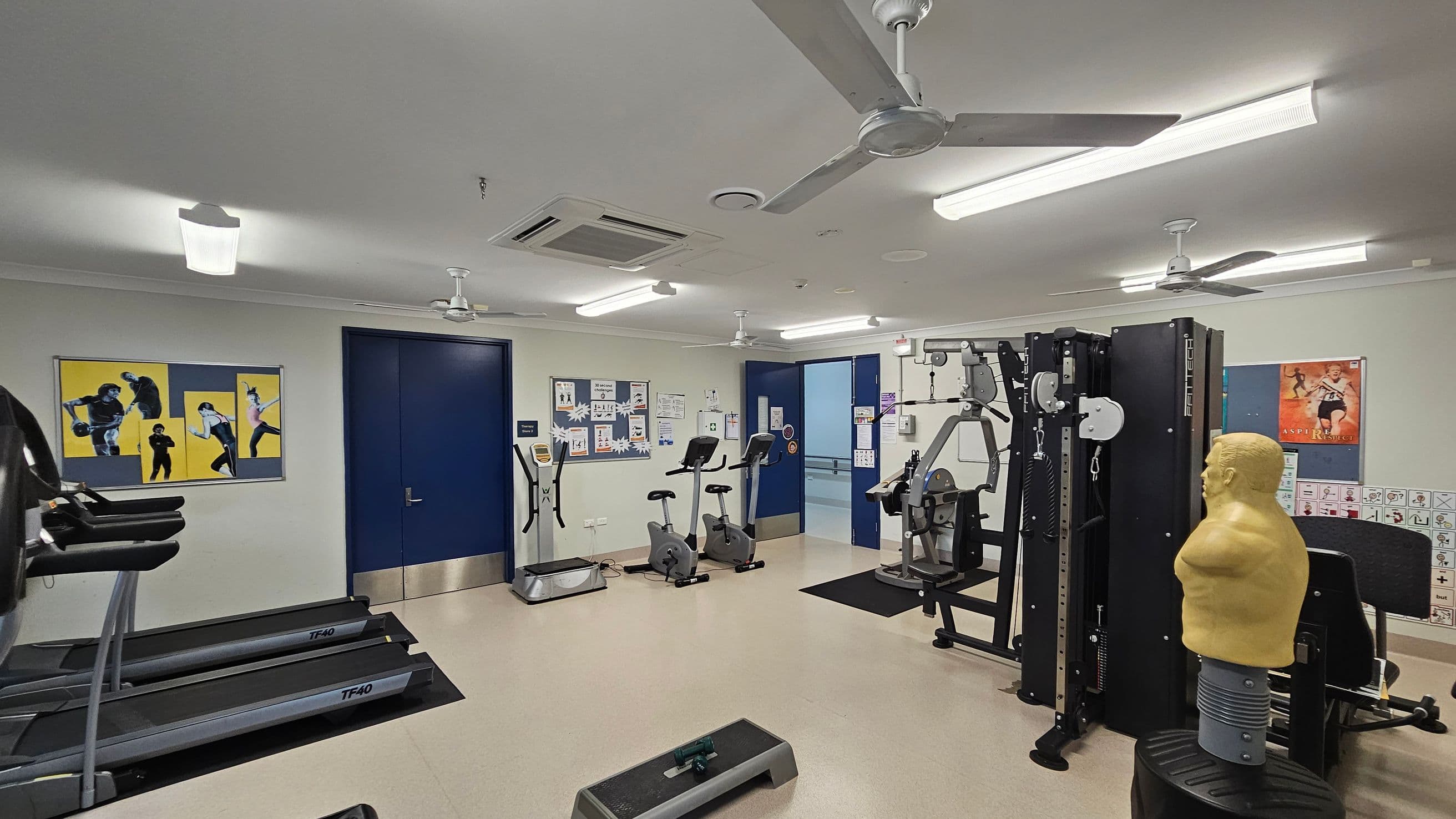 A gym room with various exercise equipment. On the left, there are treadmills and a wall with three fitness posters. In the centre, two stationary bikes and an elliptical machine are positioned near a blue door. The right side features a multi-gym machine and a punching dummy. The walls have motivational posters and informational boards. The ceiling has fluorescent lights and ceiling fans, and the floor is covered with light-coloured tiles.